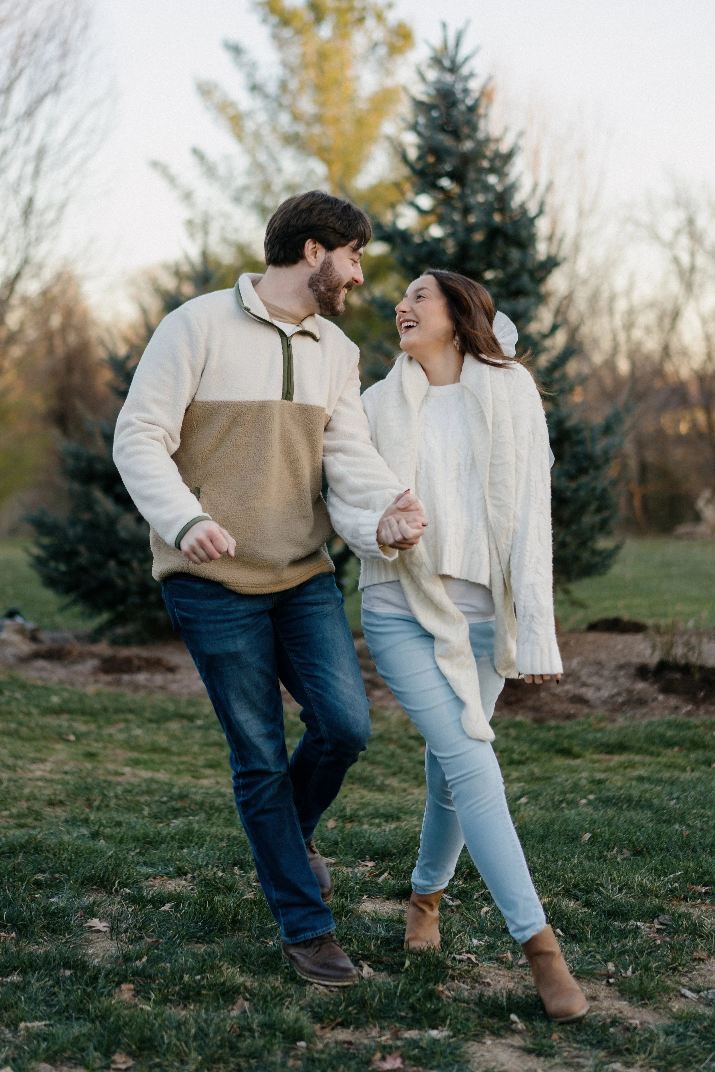 A smiling couple walking hand in hand outdoors in a park or garden during fall, surrounded by trees with autumn leaves.