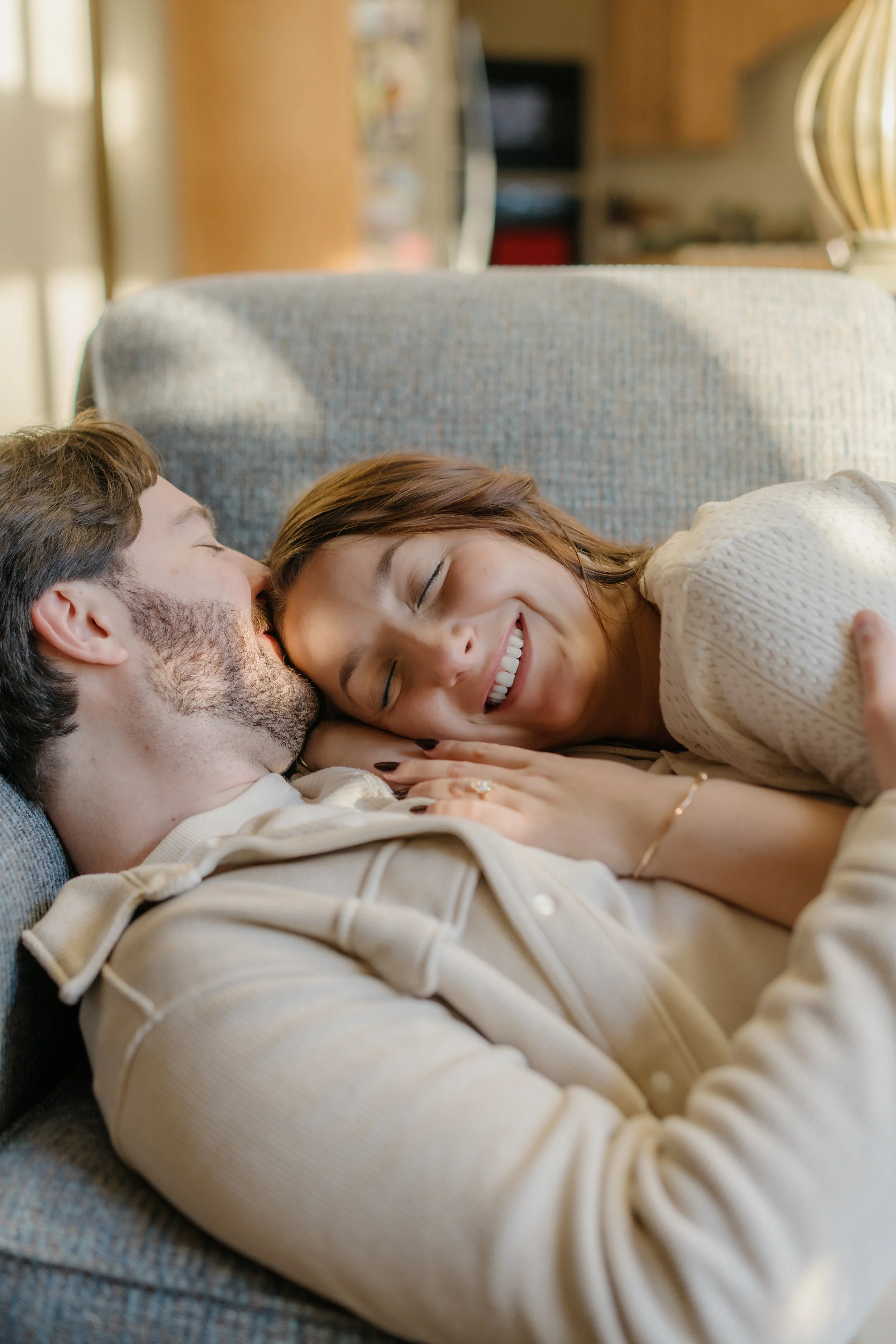 A woman and man are lying on a gray couch, smiling and cuddling. The woman is lying on her side, facing the man, with her hand resting on his chest. The man is lying on his back with his head turned toward her. They are in a cozy, warmly lit living r