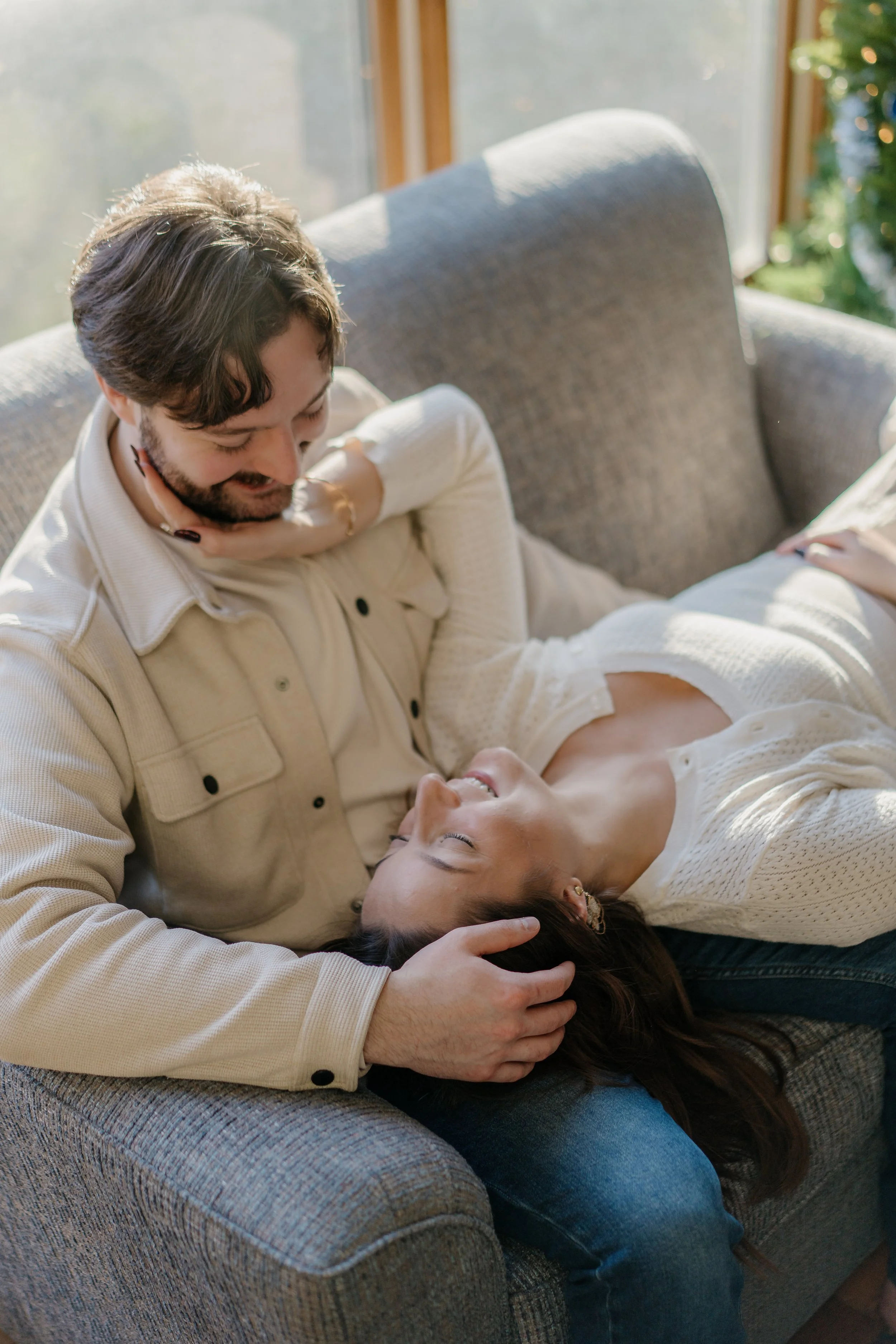 A man and woman sitting on a couch, smiling and looking at each other affectionately, with the woman lying down with her head on the man's lap.