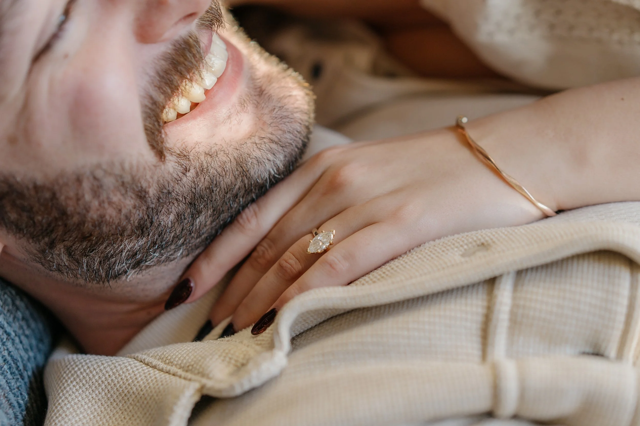Close-up of a smiling man with a beard lying down, showing his teeth, with a hand resting on his chest accessorized with a ring and bracelet, and a woman’s hand with a ring on her finger also resting on his chest.