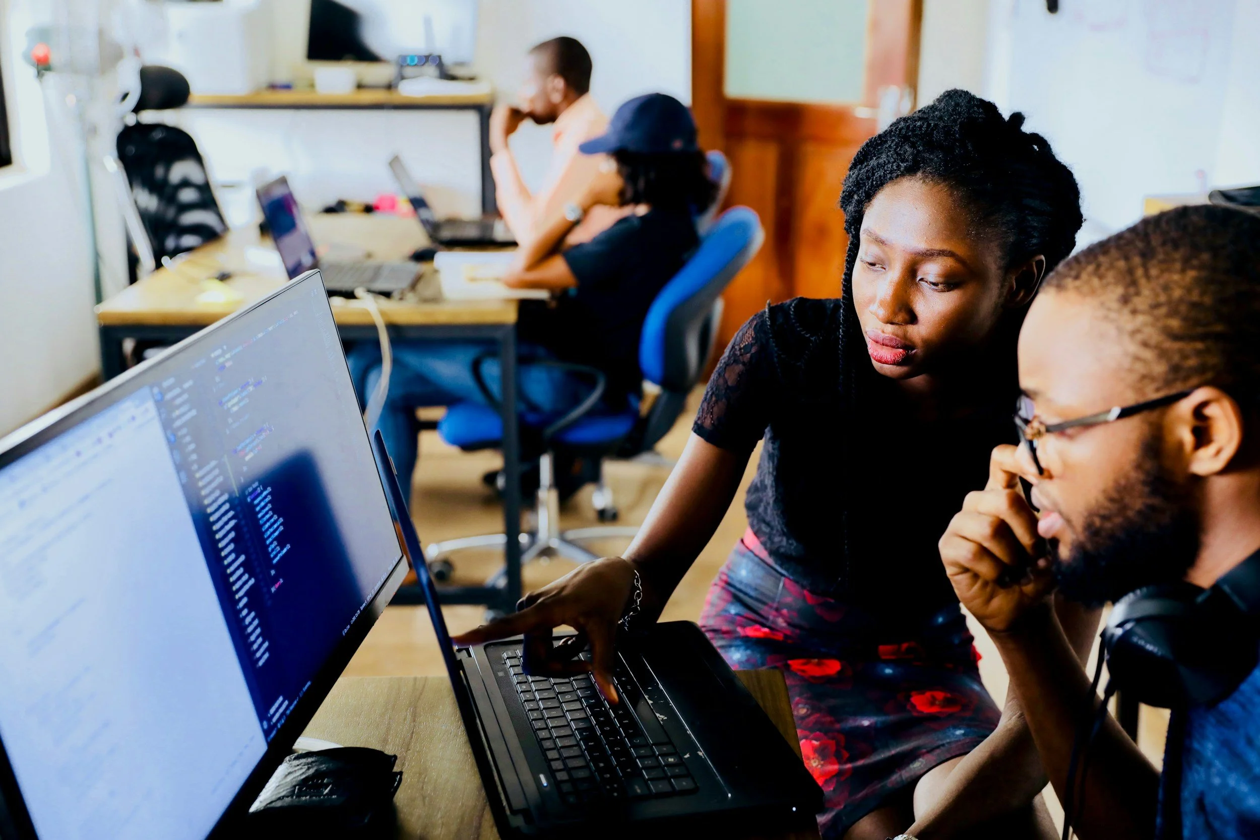 A woman and a man working on a laptop in an office, with two other people working at desks in the background.
