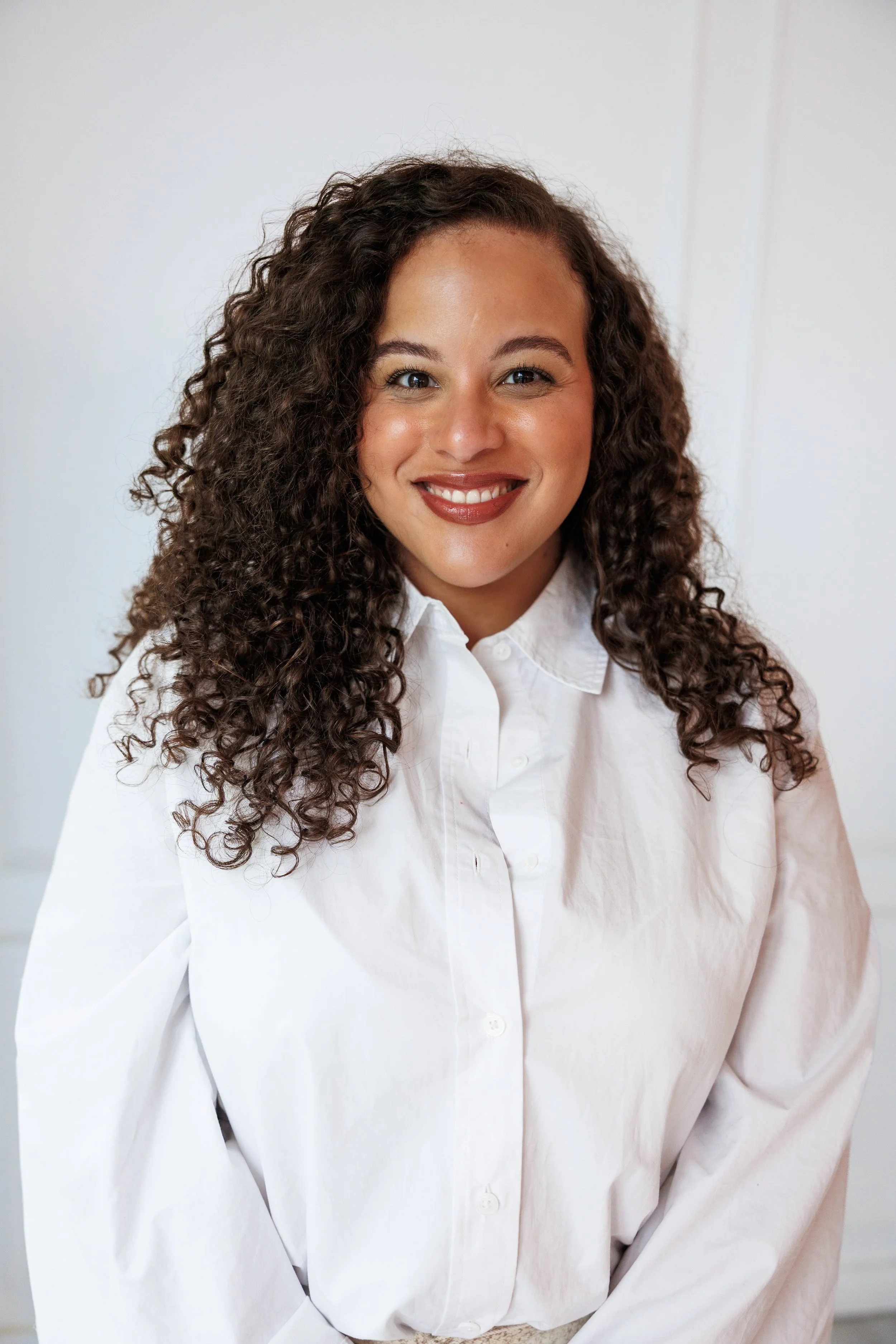 A woman with curly dark hair wearing a light purple jumpsuit stands against a purple background, looking at the camera with arms crossed.