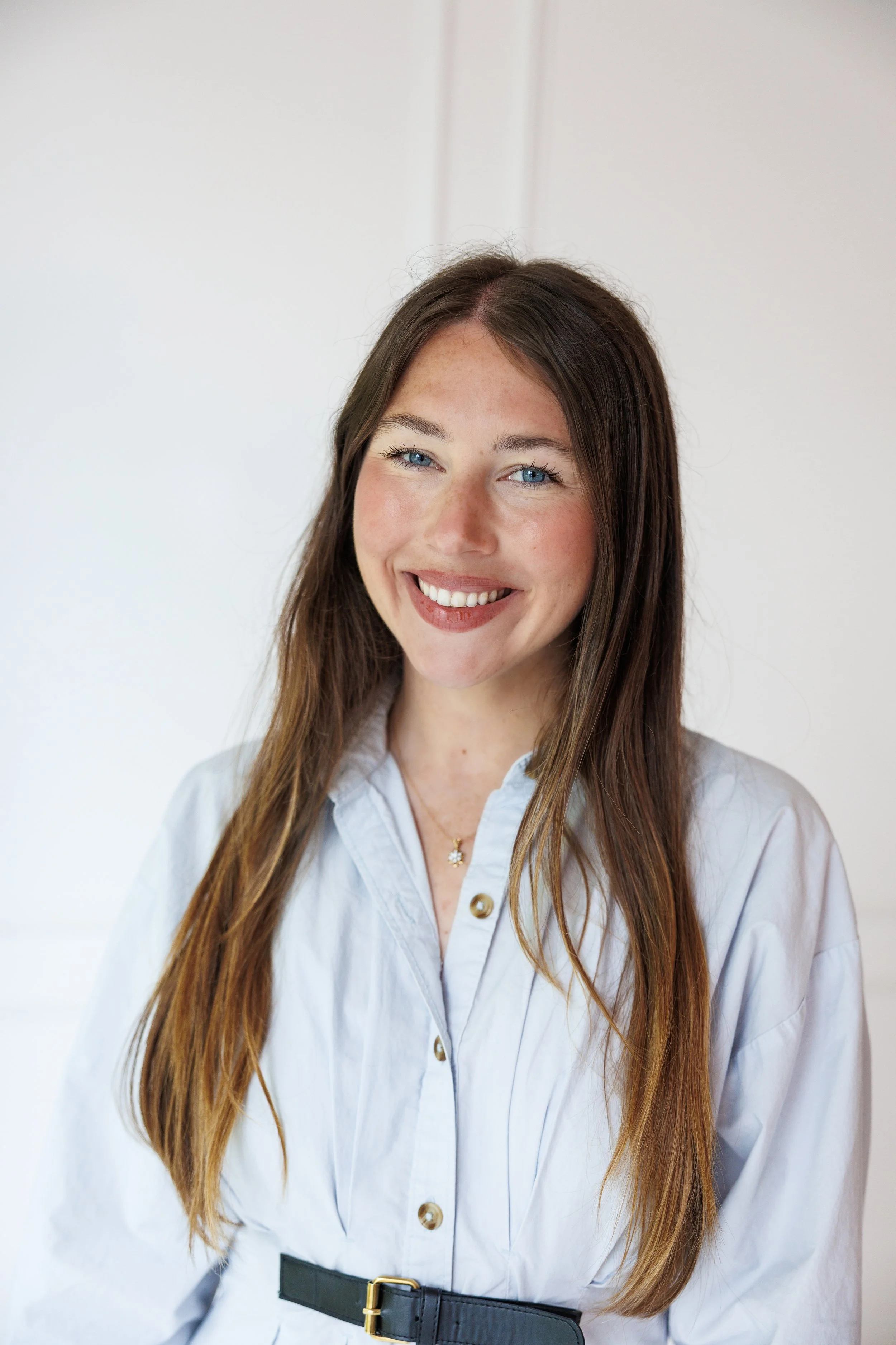A woman with long, wavy light brown hair and blue eyes smiling, wearing a black blazer and a green ruffled blouse against a gray background.