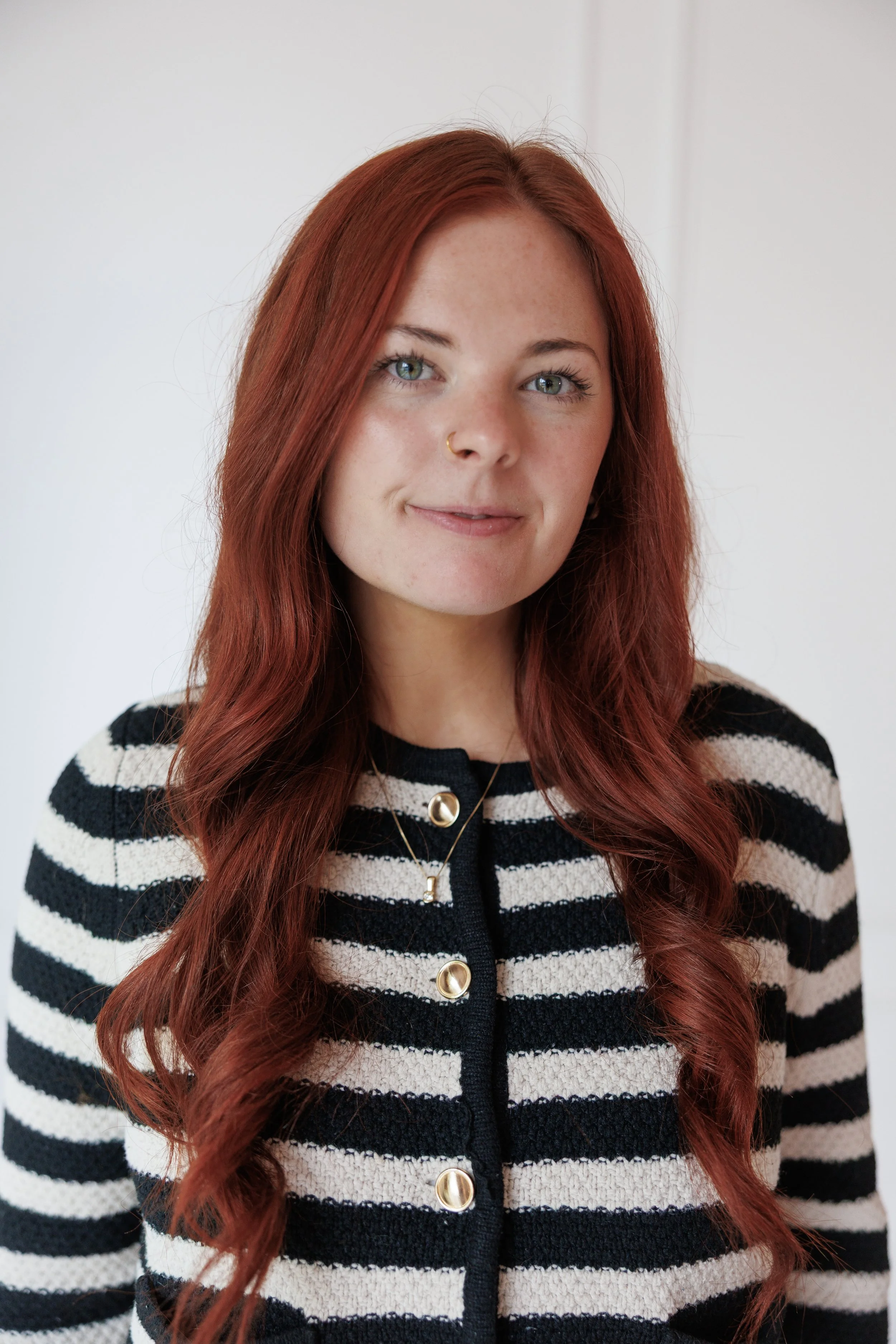 A young woman with long, red hair and fair skin smiling while standing in front of a red brick wall. She is wearing a beige blazer over a white t-shirt and a small necklace.
