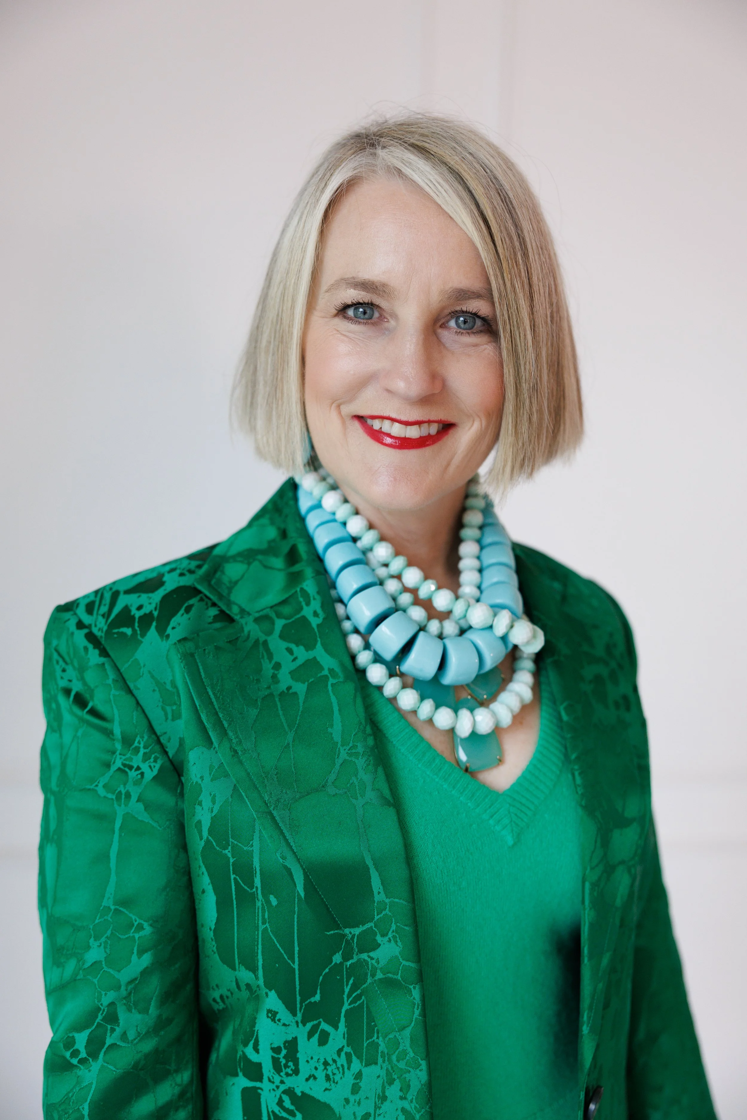 A woman with short blonde hair wearing a green blazer and a patterned shirt, smiling against a green and white geometric background.