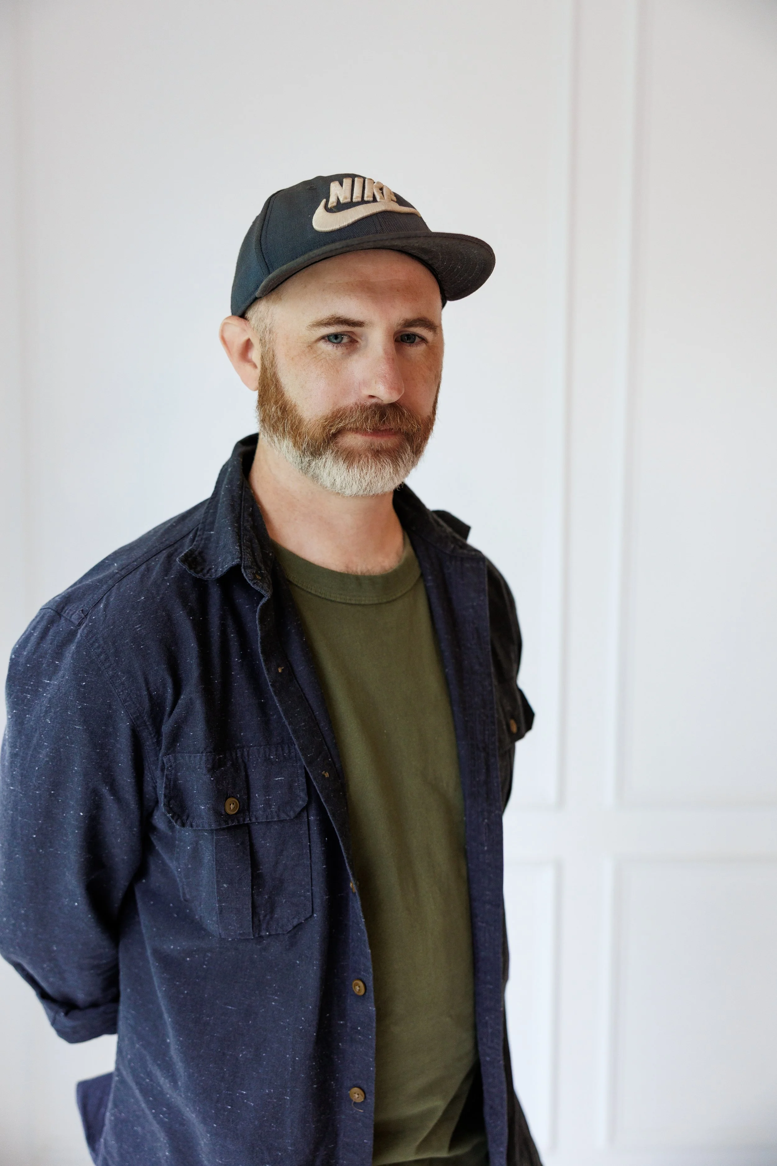 Black and white portrait of a man with a beard wearing a baseball cap and a camouflage jacket, looking to the side against a plain background.