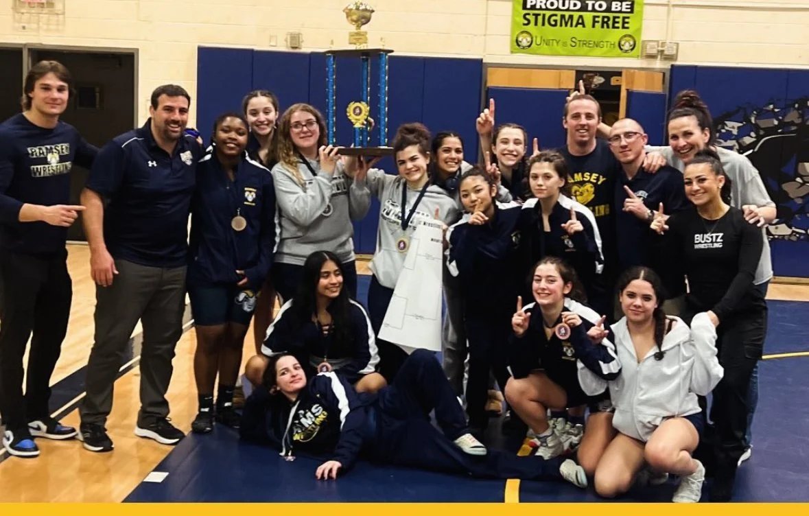 Group of eight young girls in matching navy blue and white school jackets sitting on gymnasium bleachers, smiling at the camera.