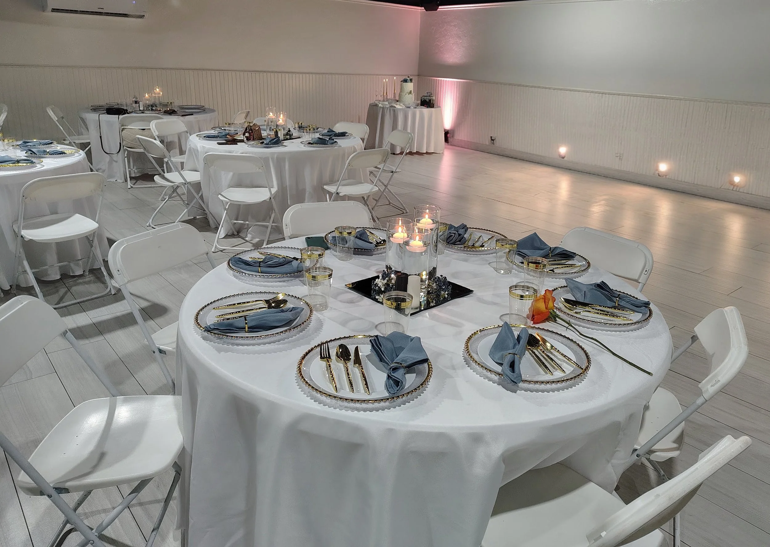 A banquet hall decorated for an event with round tables covered in white tablecloths. Each table has a place setting with dark blue napkins, gold cutlery, and clear glasses. Centerpieces with candles are on the tables. In the background, there is a table with a white tablecloth, topped with a tiered dessert stand and a cake. The walls are plain, and the floor is light-colored wood.