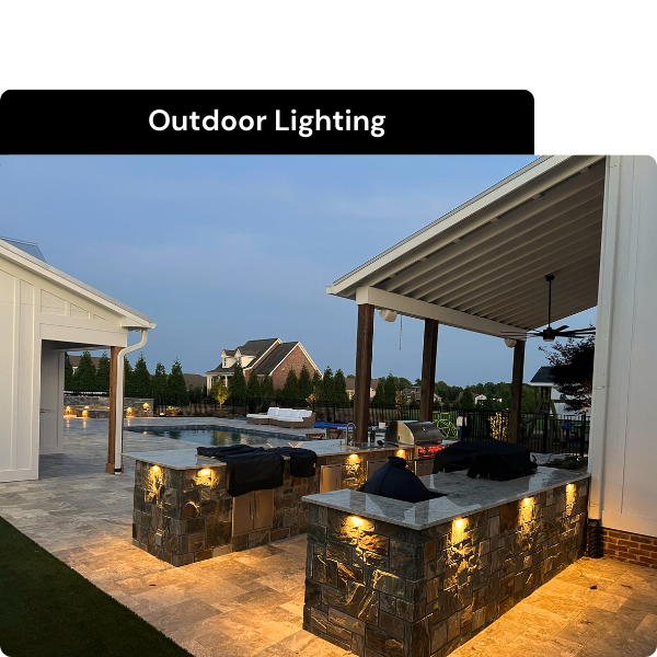 Outdoor kitchen with stone counters and lighting, covered patio, swimming pool, and neighboring houses in the background during evening. Installed by B & C Designs in the Triangle, North Carolina.