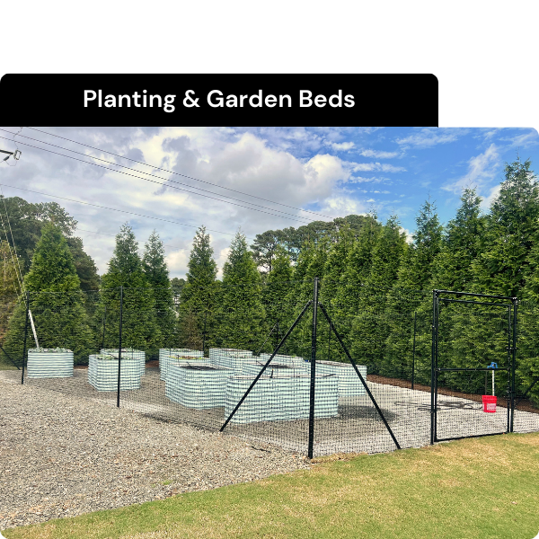 Garden beds made of stacked white cinder blocks, enclosed by black metal fencing, with a sign that says 'Planting & Garden Beds' and a background of green trees under a partly cloudy sky. Installed by B & C Designs in the Triangle, North Carolina.