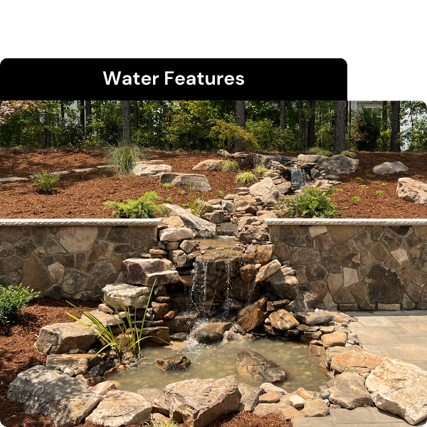 Garden water feature with rocks, small waterfall, and pond, surrounded by mulch and plants, with a stone wall background and trees. Installed by B & C Designs in the Triangle, North Carolina.