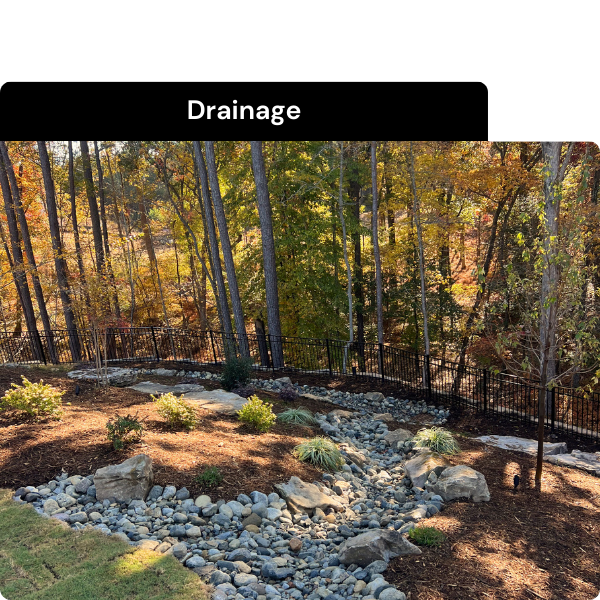 A landscaped yard with a dry creek bed made of river rocks, surrounded by plants and a black metal fence, with trees and autumn foliage in the background. Installed by B & C Designs in the Triangle, North Carolina.