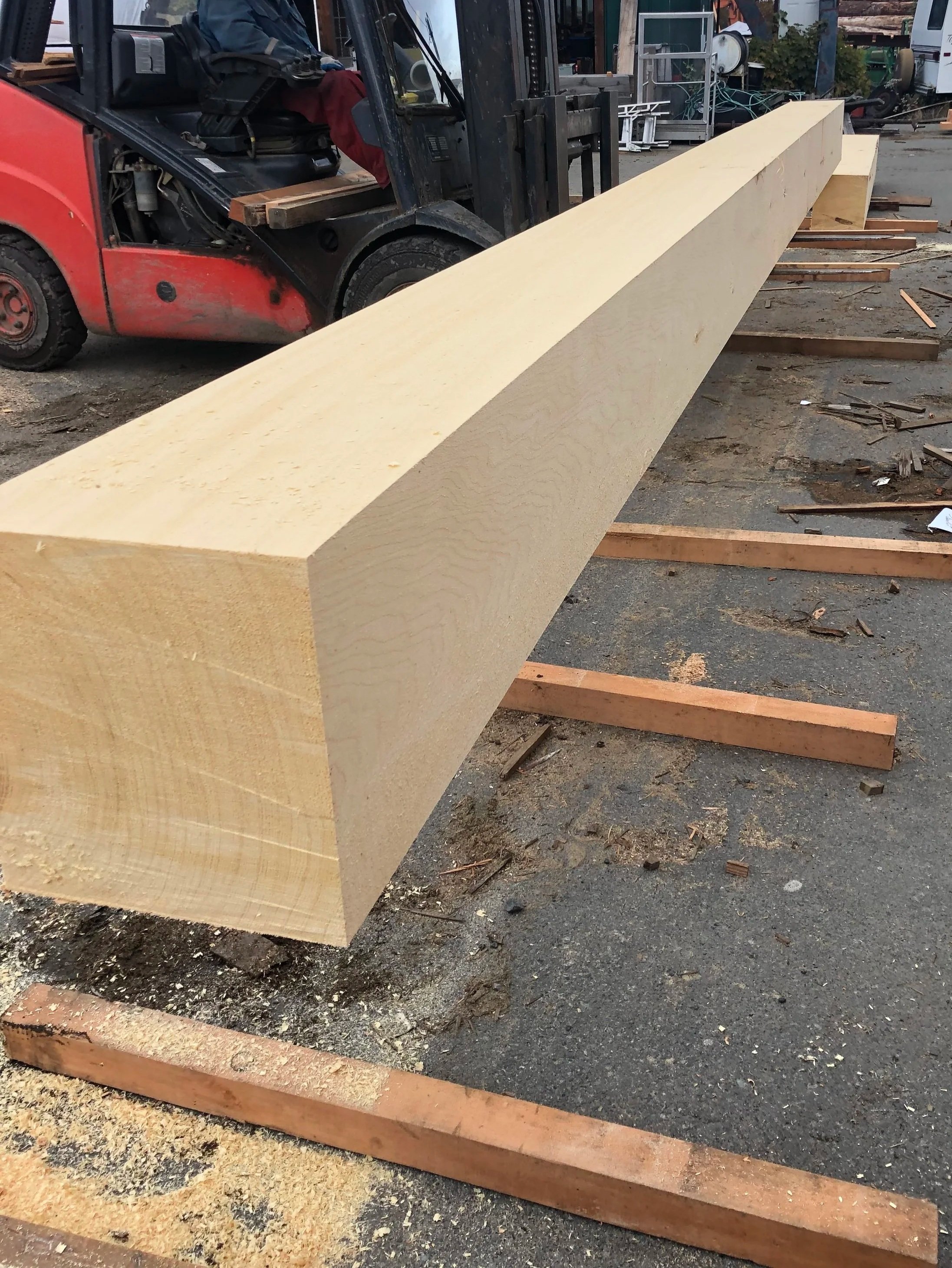 A large, unfinished wooden beam resting on a gravel ground in a construction yard, with wood shavings and scattered sawdust around.