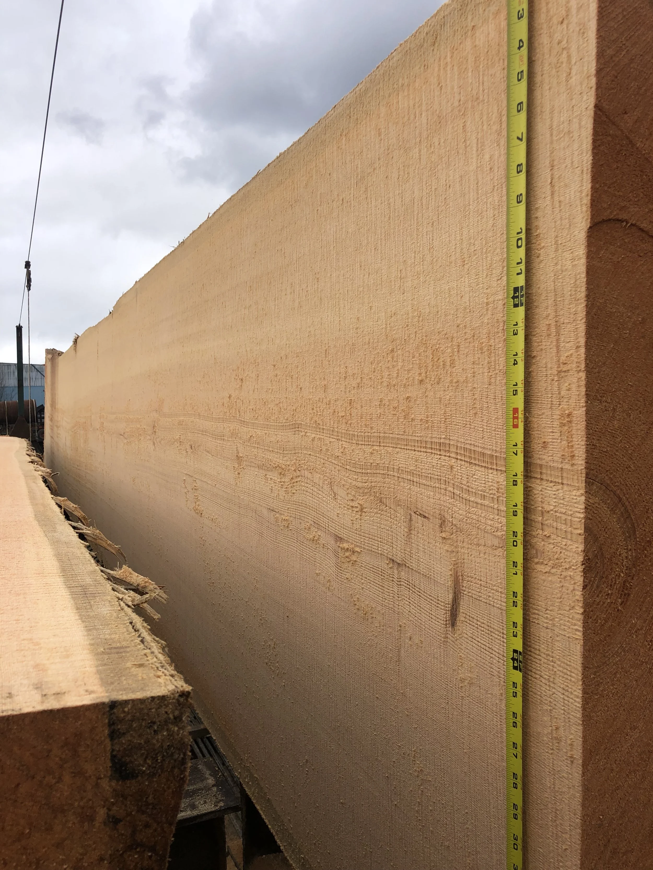 A large, freshly cut wooden plank with a measuring tape along its length. The background shows an overcast sky and some industrial equipment.