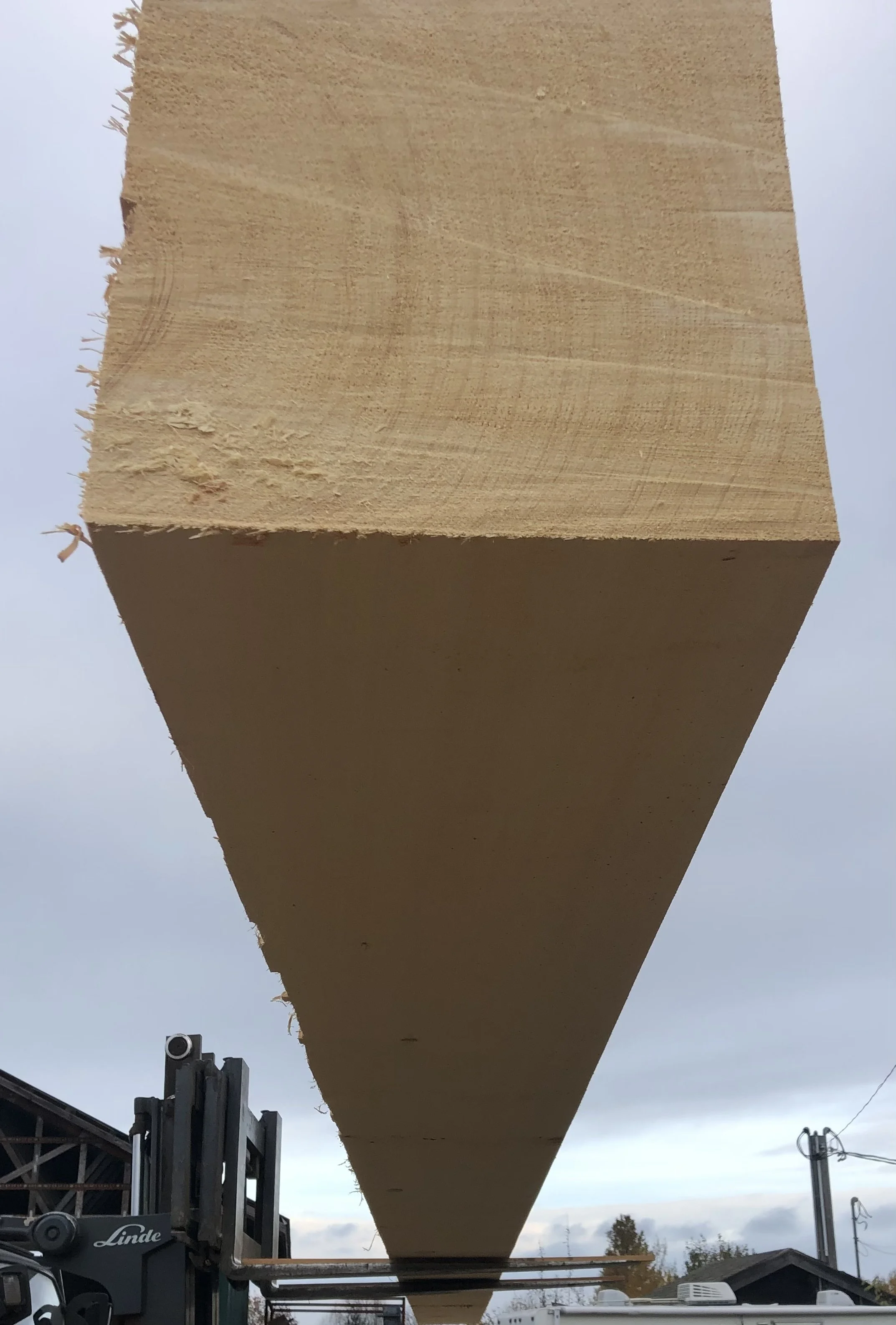 Construction site with a large wooden beam being positioned by a forklift, under cloudy sky.