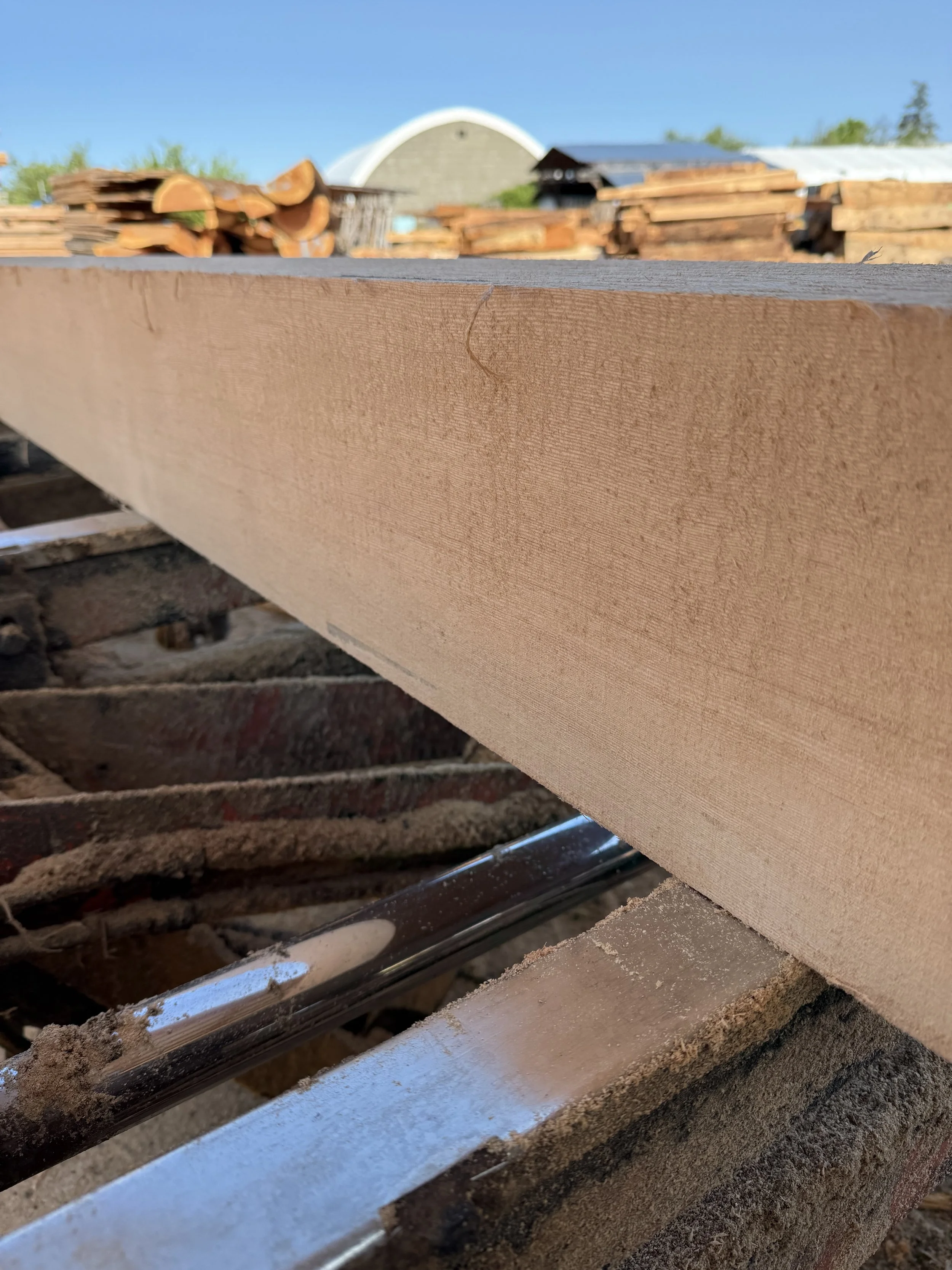 Close-up of a piece of light-colored wood on a saw horse with a background of stacked wood pallets and a building with a curved roof under a clear blue sky.