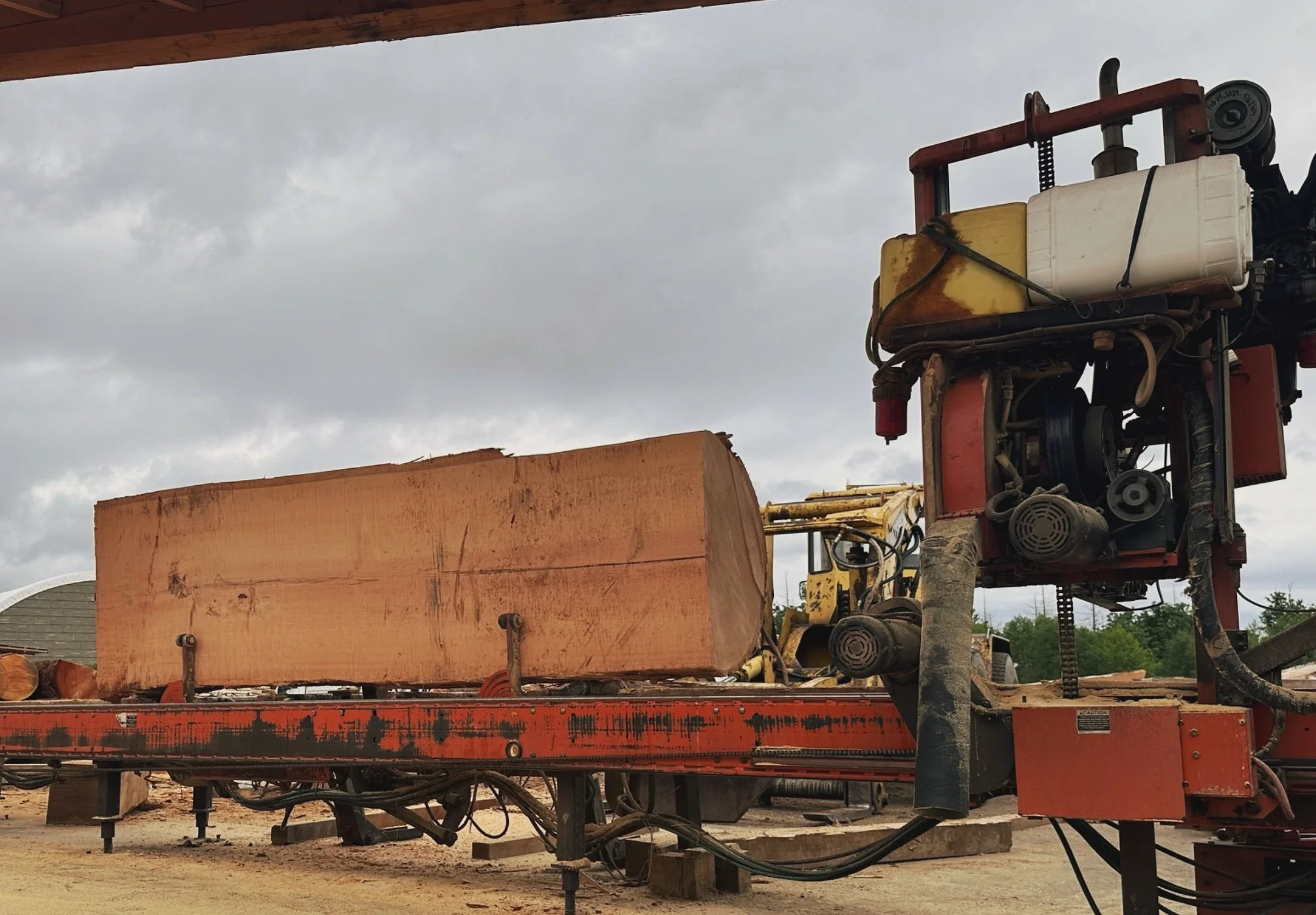 A large orange sawmill machine cutting a thick log of wood outdoors, with a cloudy sky overhead.