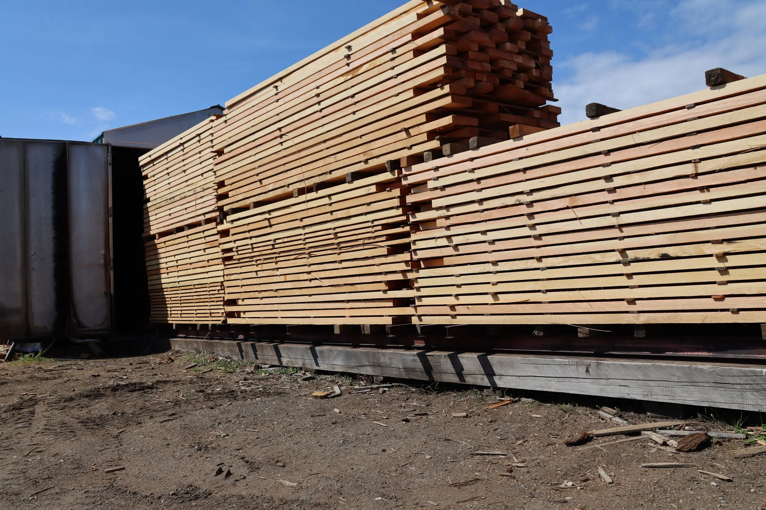 Large stack of wooden planks on a flatbed truck outdoors against a blue sky.