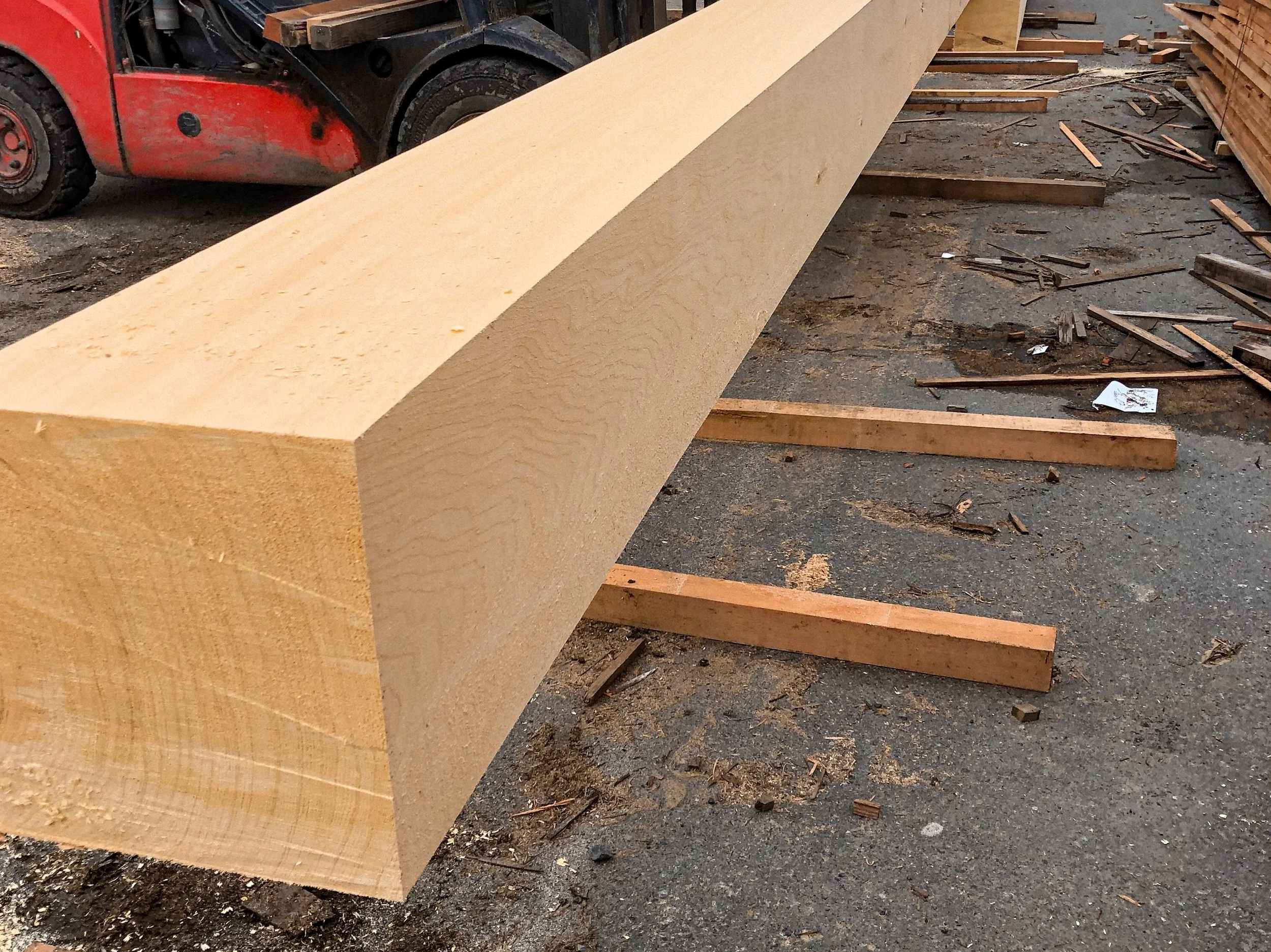 A large wooden beam placed on wooden supports in a workshop with sawdust and small wood pieces on the ground.