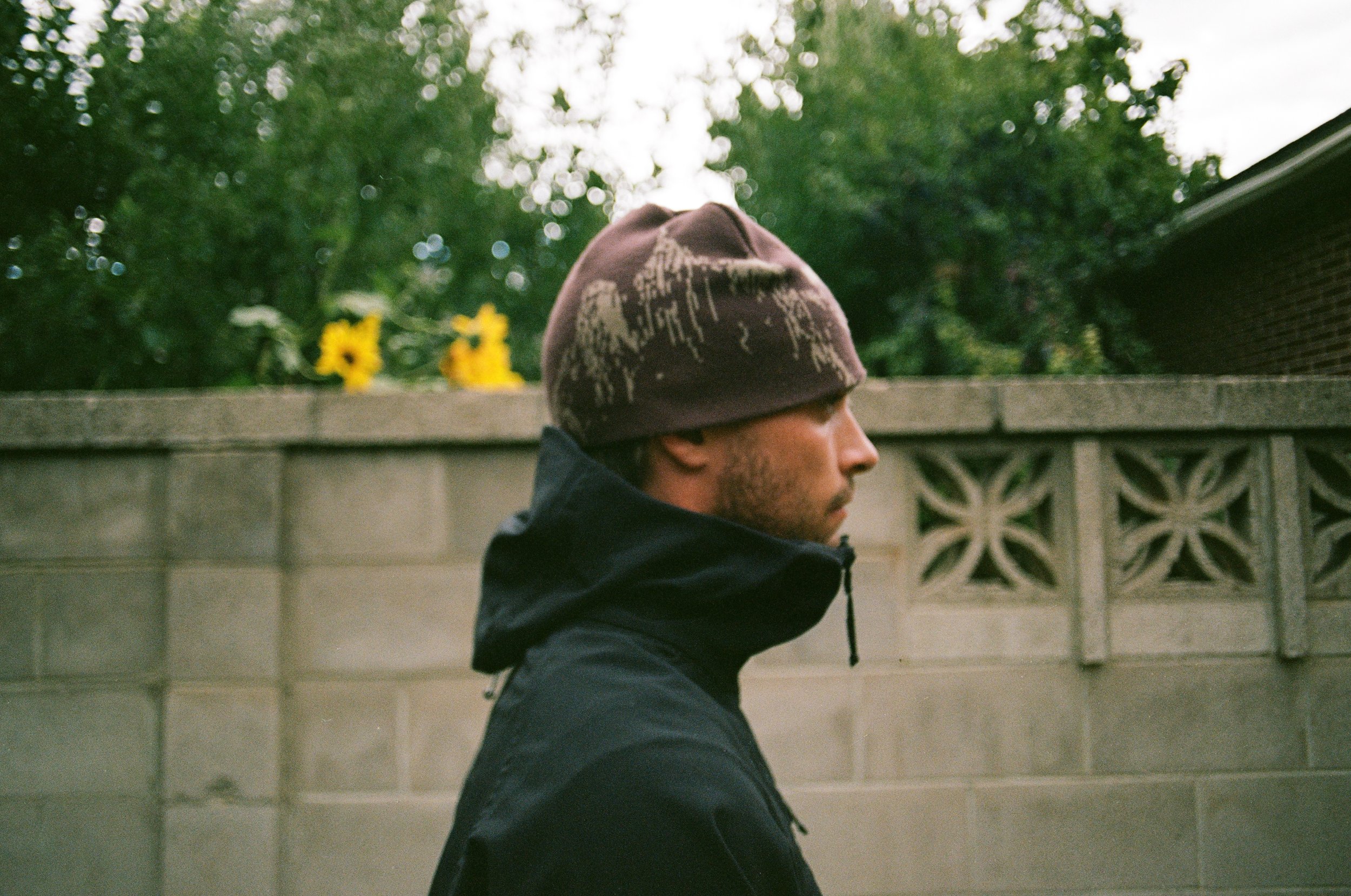 Person wearing a brown beanie and a dark jacket, walking in front of a patterned concrete wall with sunflowers in the background.