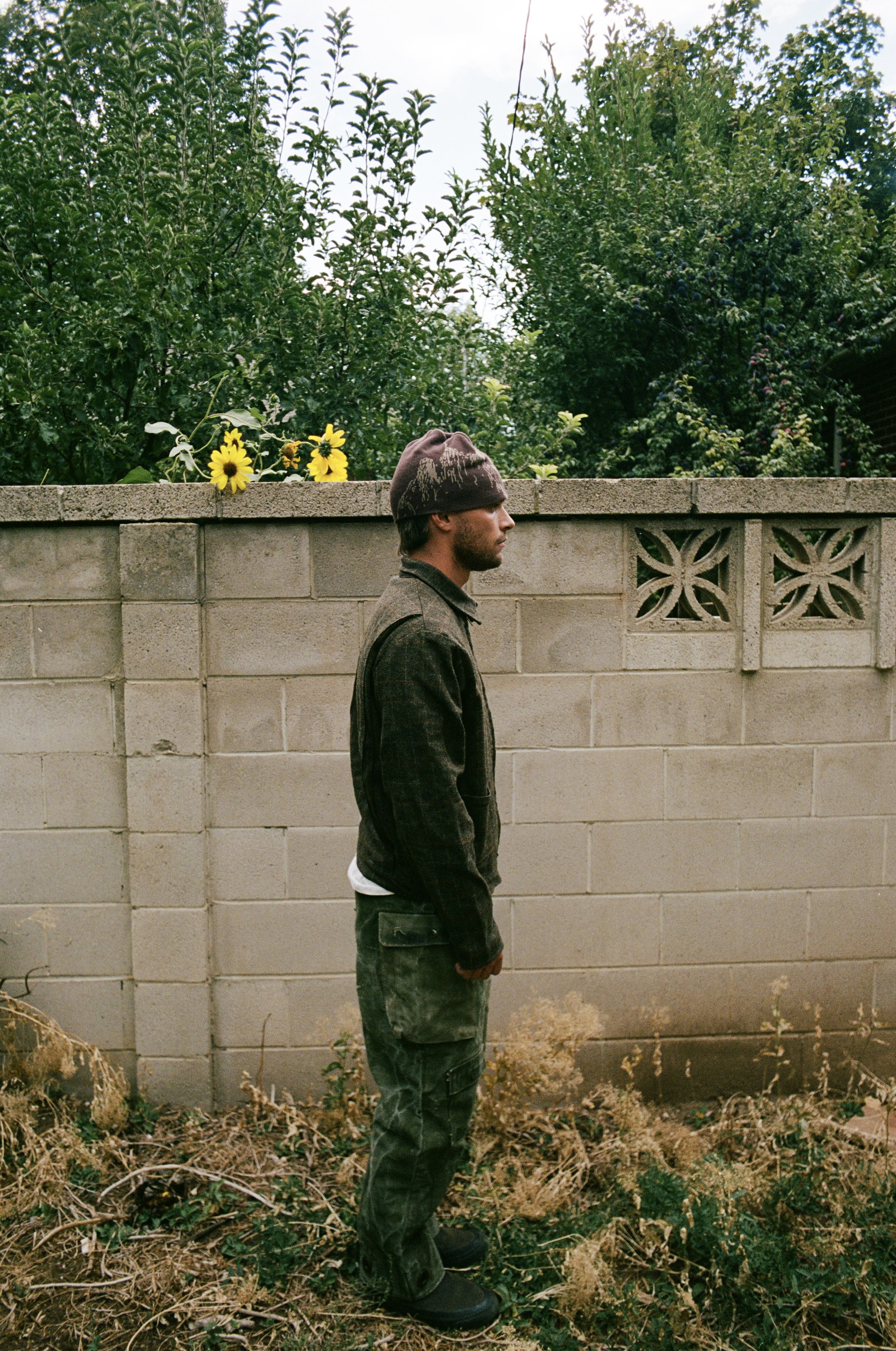 Man in brown jacket and beanie standing beside a concrete wall, with sunflowers and bushes in the background.