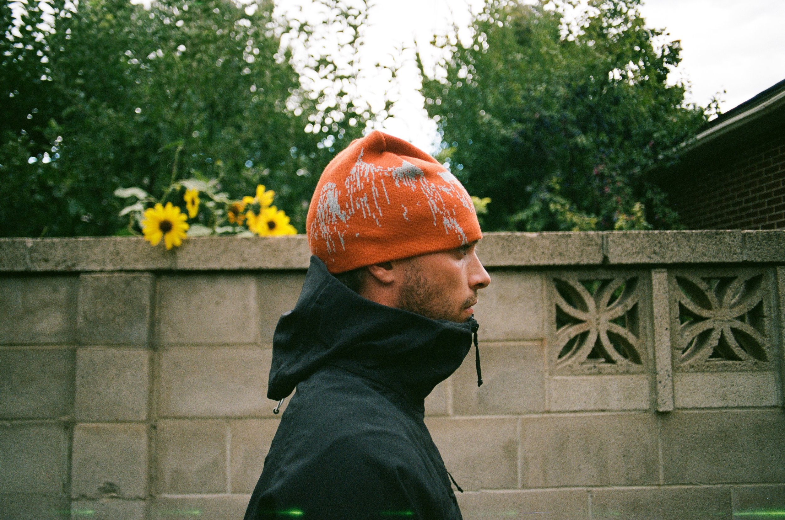 Man in profile wearing an orange beanie and dark jacket, standing in front of a stone wall with sunflowers and greenery in the background.