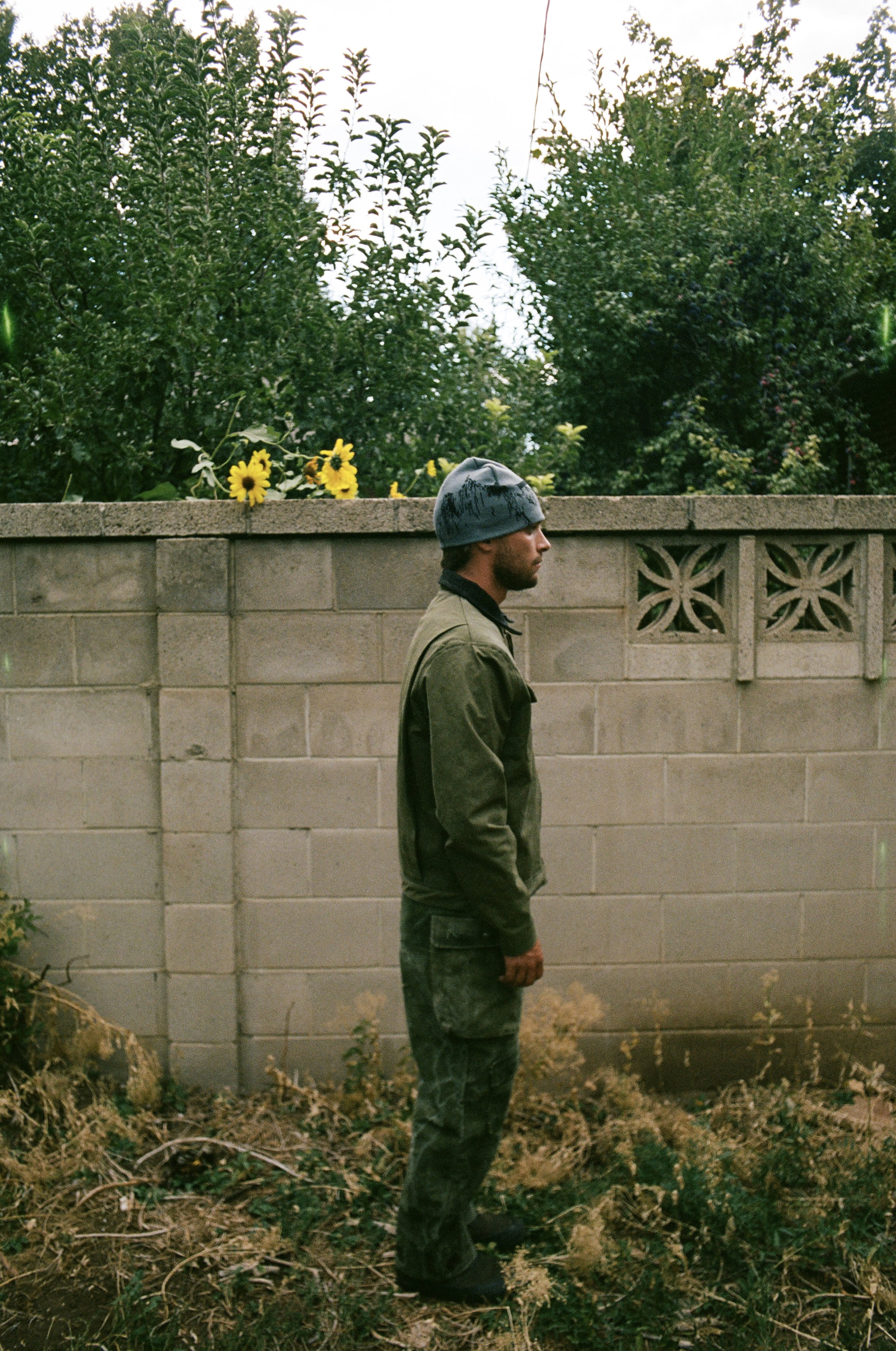 Man in a beanie and green jacket standing beside a concrete wall with sunflowers and greenery in the background.