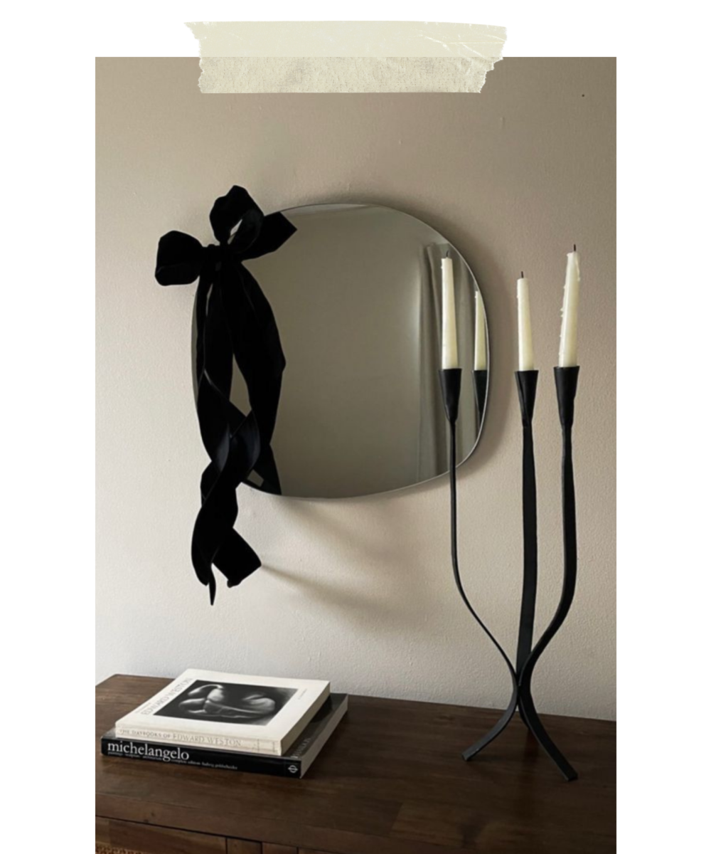 A round mirror with a black ribbon bow hanging on a white wall, a three-candle black metal candle holder with white candles, and a stack of art books on a wooden surface.