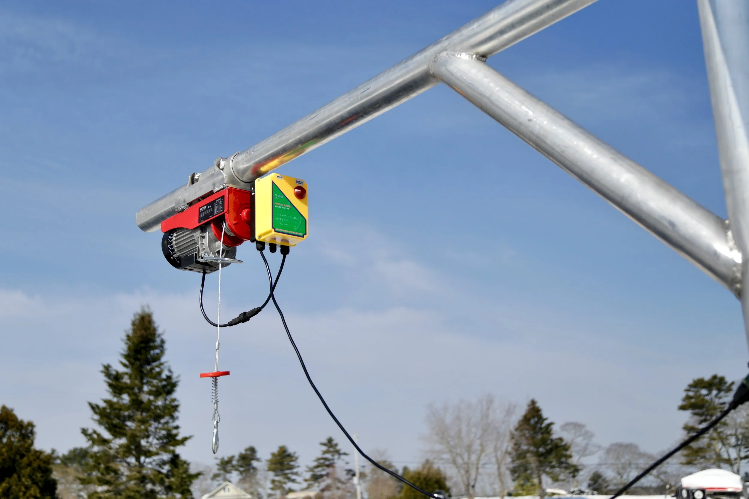Close-up of a metal davit arm with an electric hoist and hook, mounted on an aluminum frame against a blue sky with trees in the background.