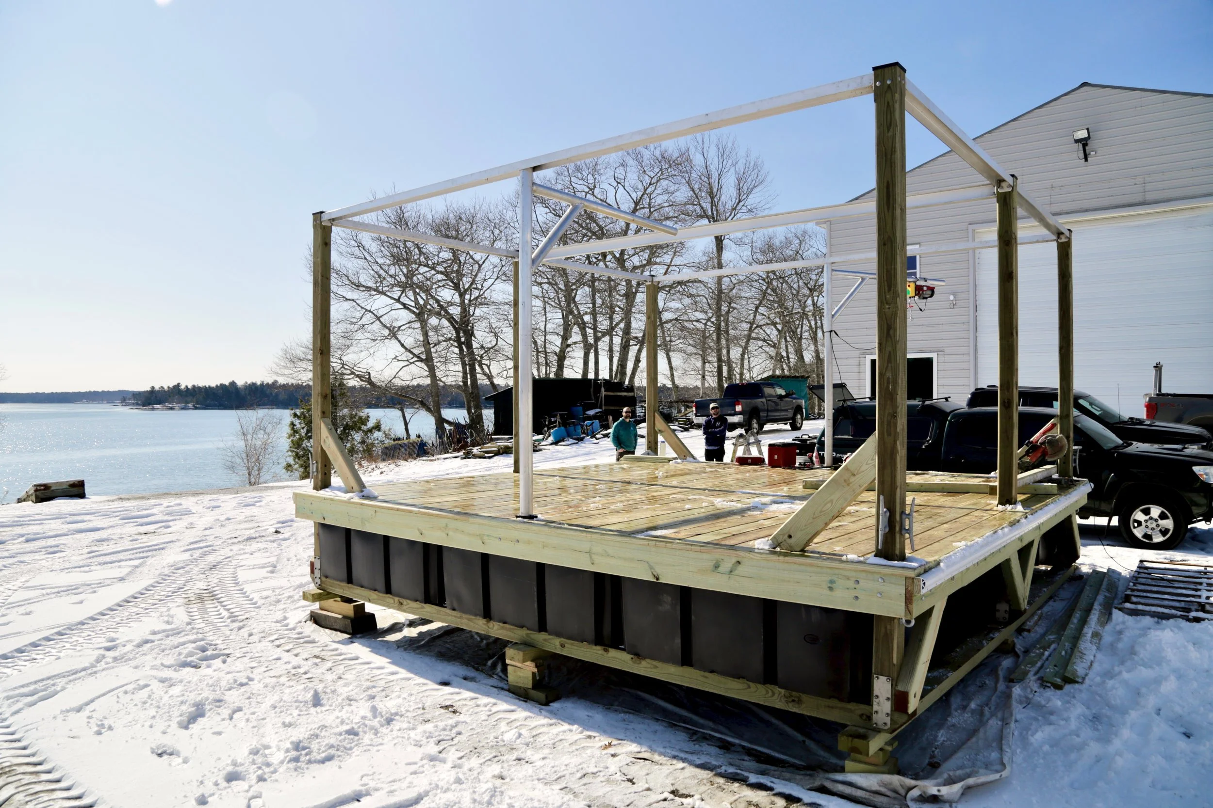 A 20 by 20 foot wooden oyster nursery float under construction at a snowy boatyard overlooking coastal water, with a metal frame structure and workers in the background.