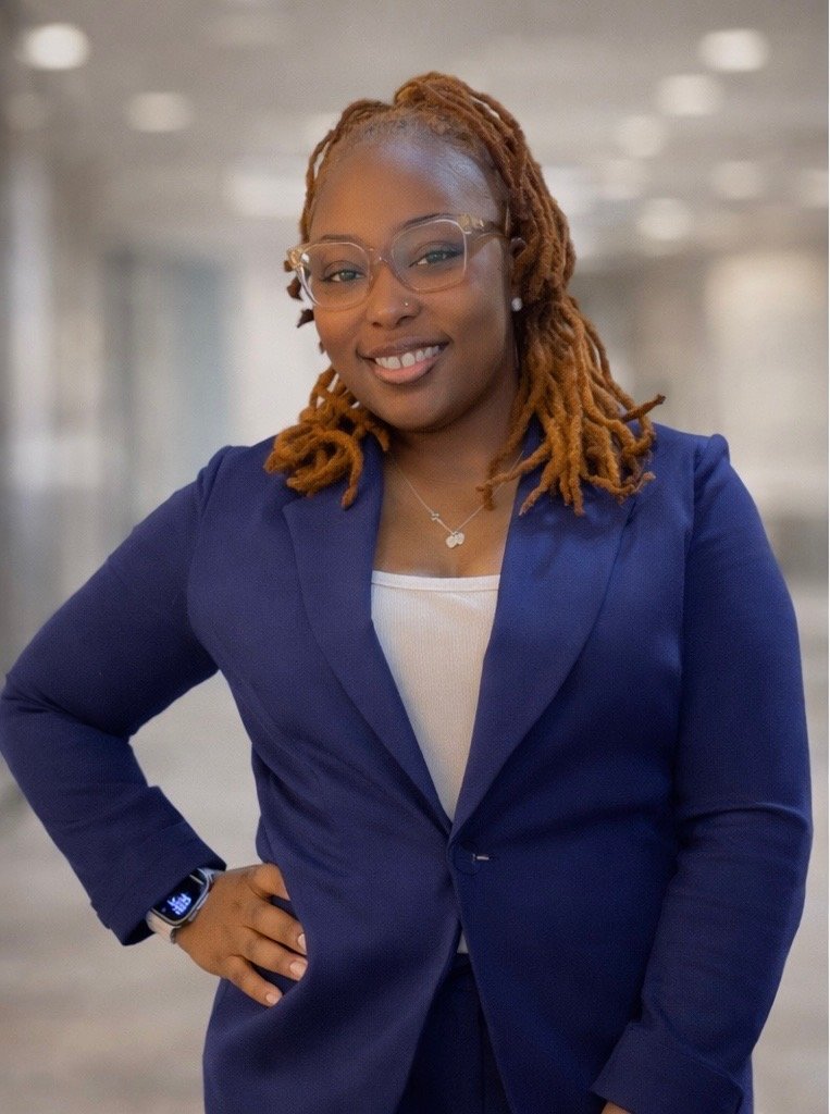 A woman with shoulder-length curly reddish-brown hair, wearing glasses, a blue blazer, and a white top, smiling confidently with her hand on her hip, in an indoor setting with blurred lighting in the background.