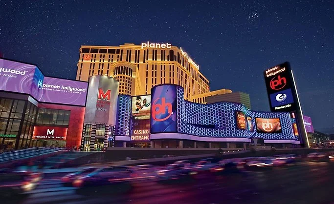 Exterior view of Planet Hollywood Resort & Casino in Las Vegas at night, featuring bright neon lights, digital billboards, and the iconic building structure.