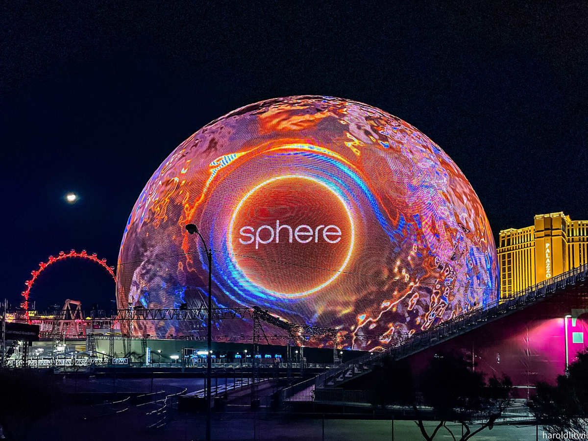 The image shows a large, illuminated dome labeled "Sphere" with colorful, dynamic lighting patterns. The setting is nighttime, with surrounding structures partially visible, including a Ferris wheel and a building labeled "Palazzo."