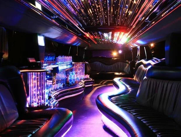 Interior of a luxury limousine with colorful LED lighting, plush seating, and a bar with drinks.