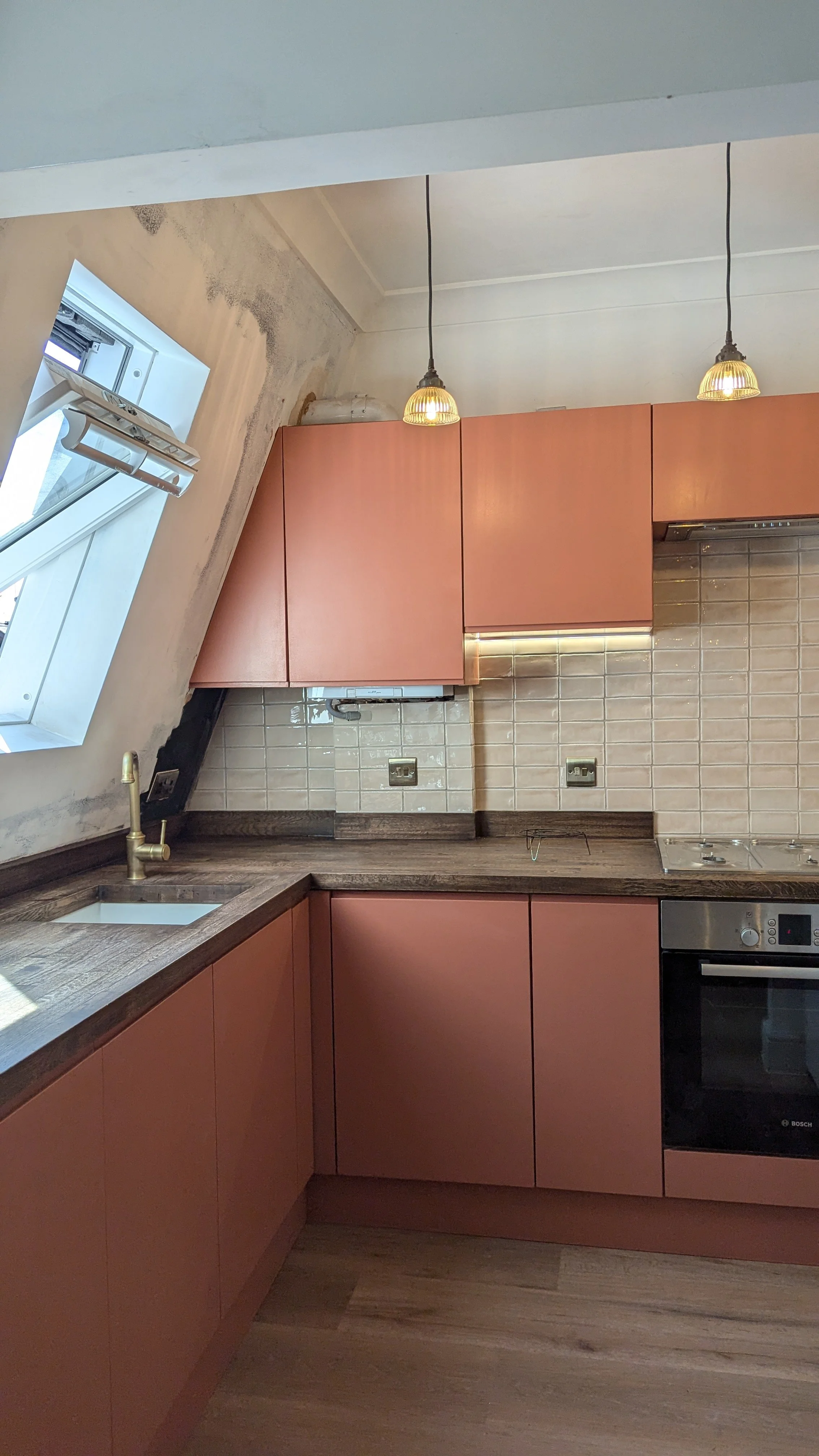 Kitchen with sloped ceiling, pink cabinets, wooden countertop, tiled backsplash, small sink, built-in oven, and two hanging pendant lights.