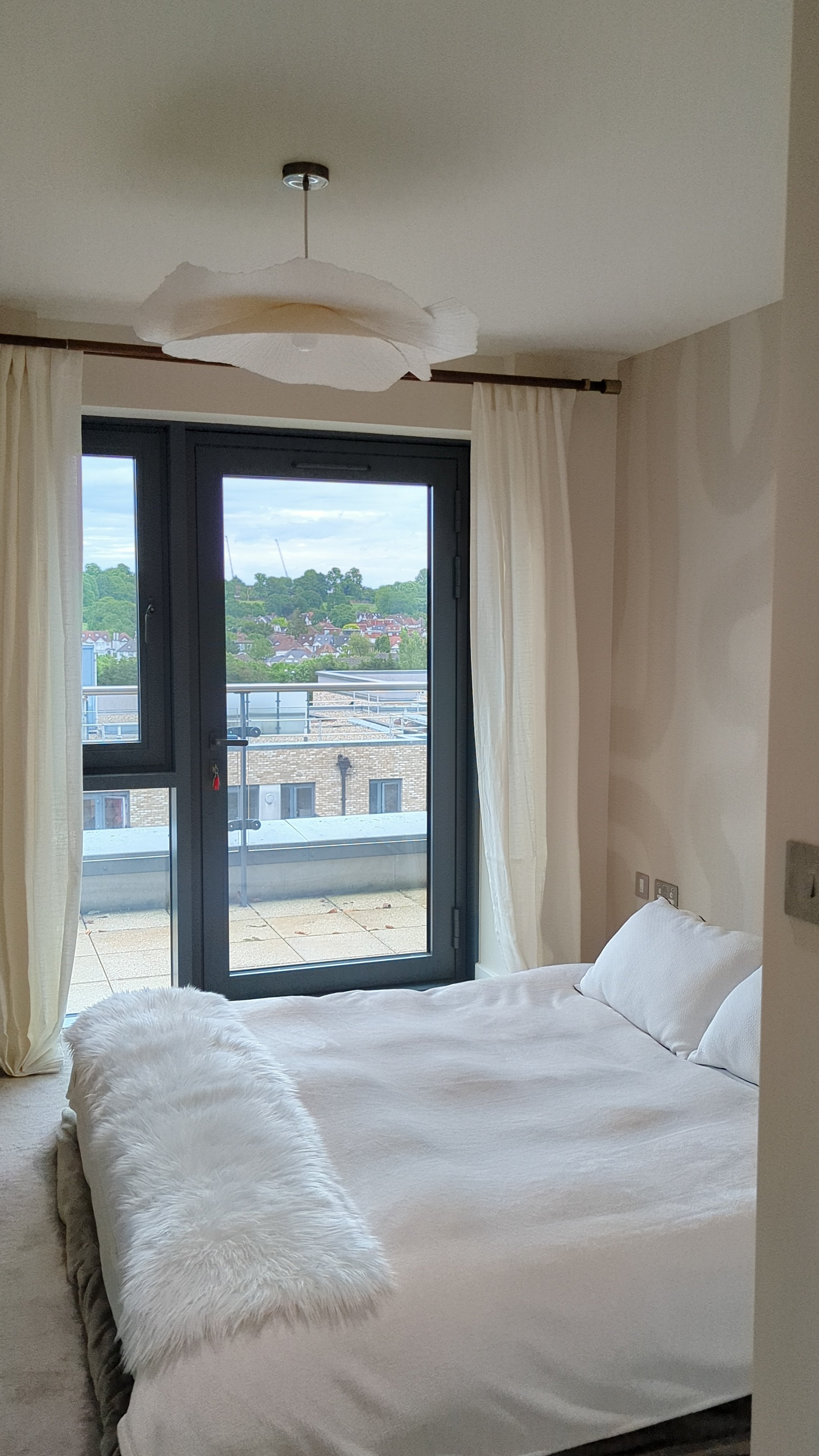 A bedroom with a bed covered with a white duvet and a white fluffy blanket, beige carpets, cream curtains, a sliding glass door leading to a balcony, and a modern white ceiling light fixture.