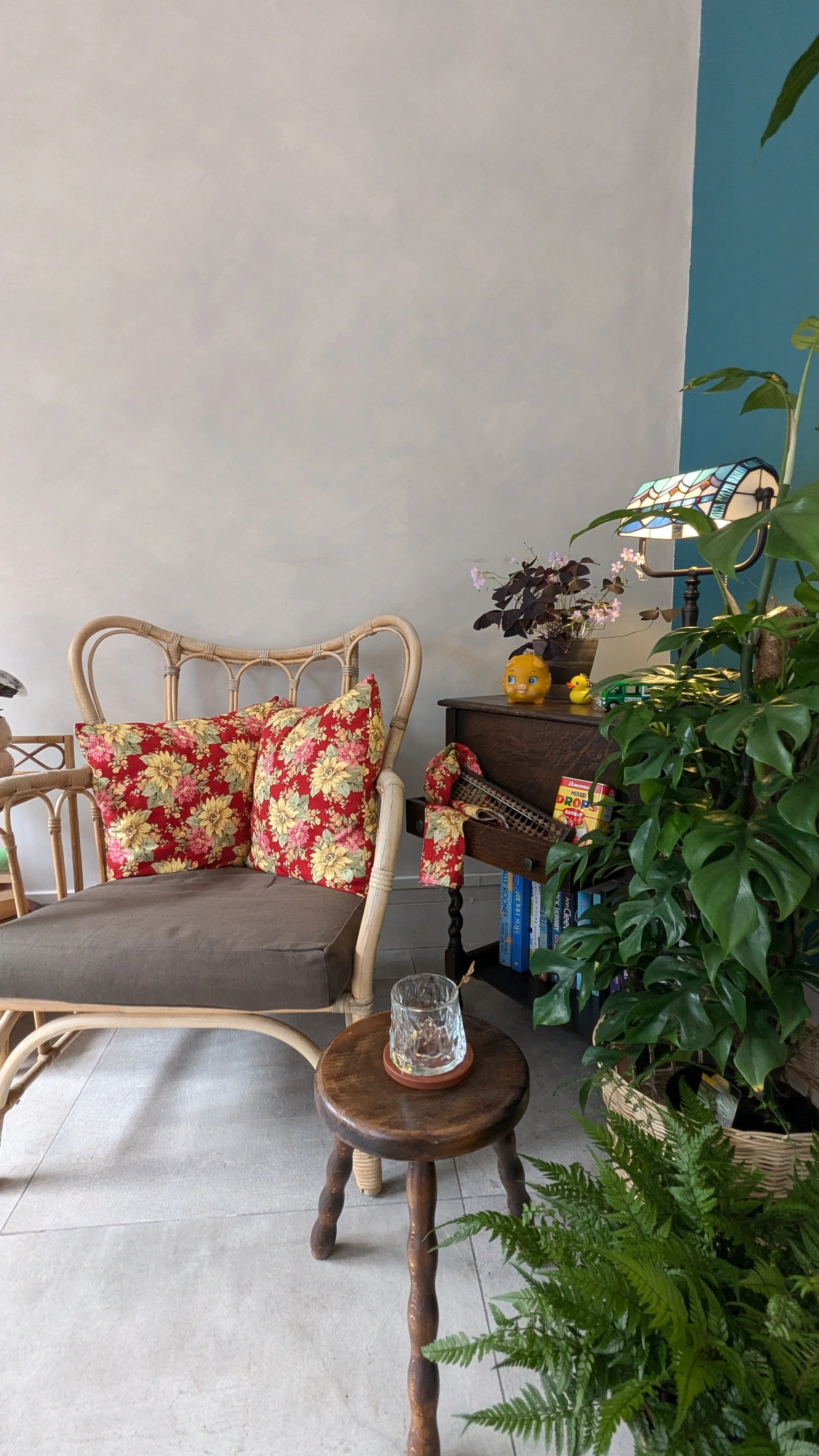 A cozy indoor nook featuring a wicker chair with floral cushions, a wooden side table with decorative items, plants, books, and a stained-glass lamp.