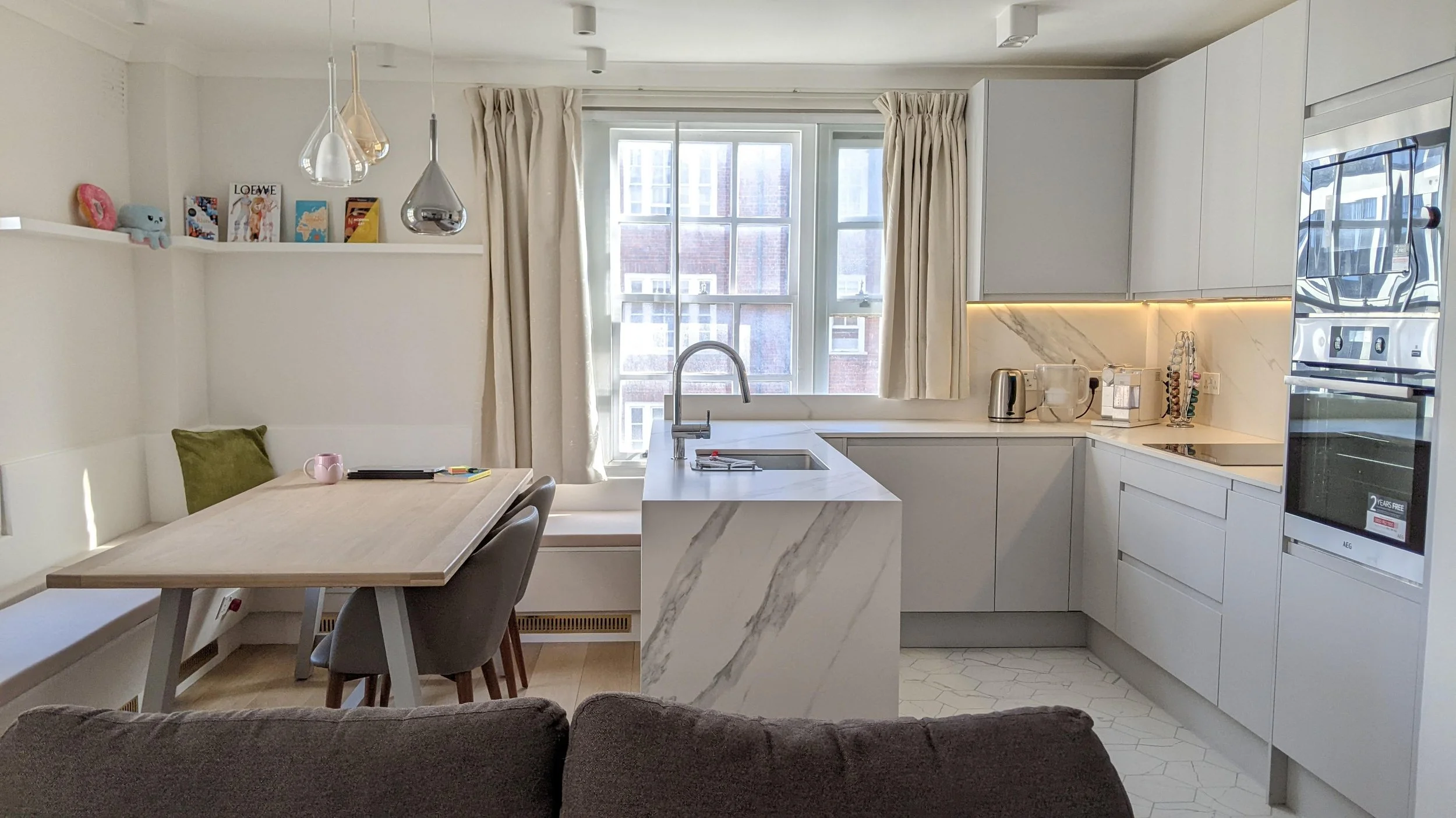 Modern kitchen with white cabinets, marble countertops, and a window with beige curtains. A dining table with chairs is visible, and there are decorative pendant lights hanging from the ceiling.