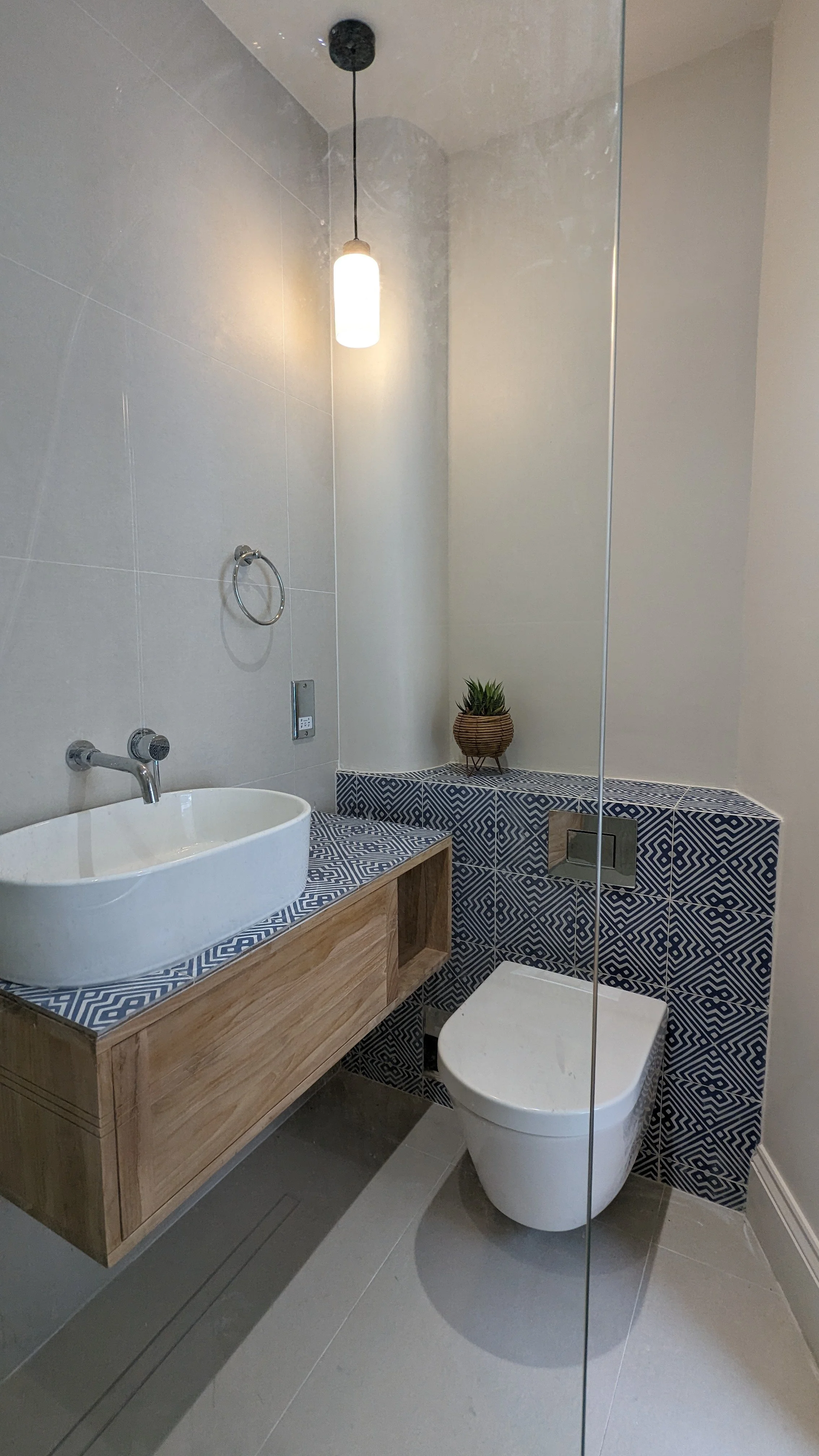 Modern bathroom with a vessel sink, wall-mounted faucet, wall light, decorative tile, small potted plant, and a toilet, separated by glass partition.