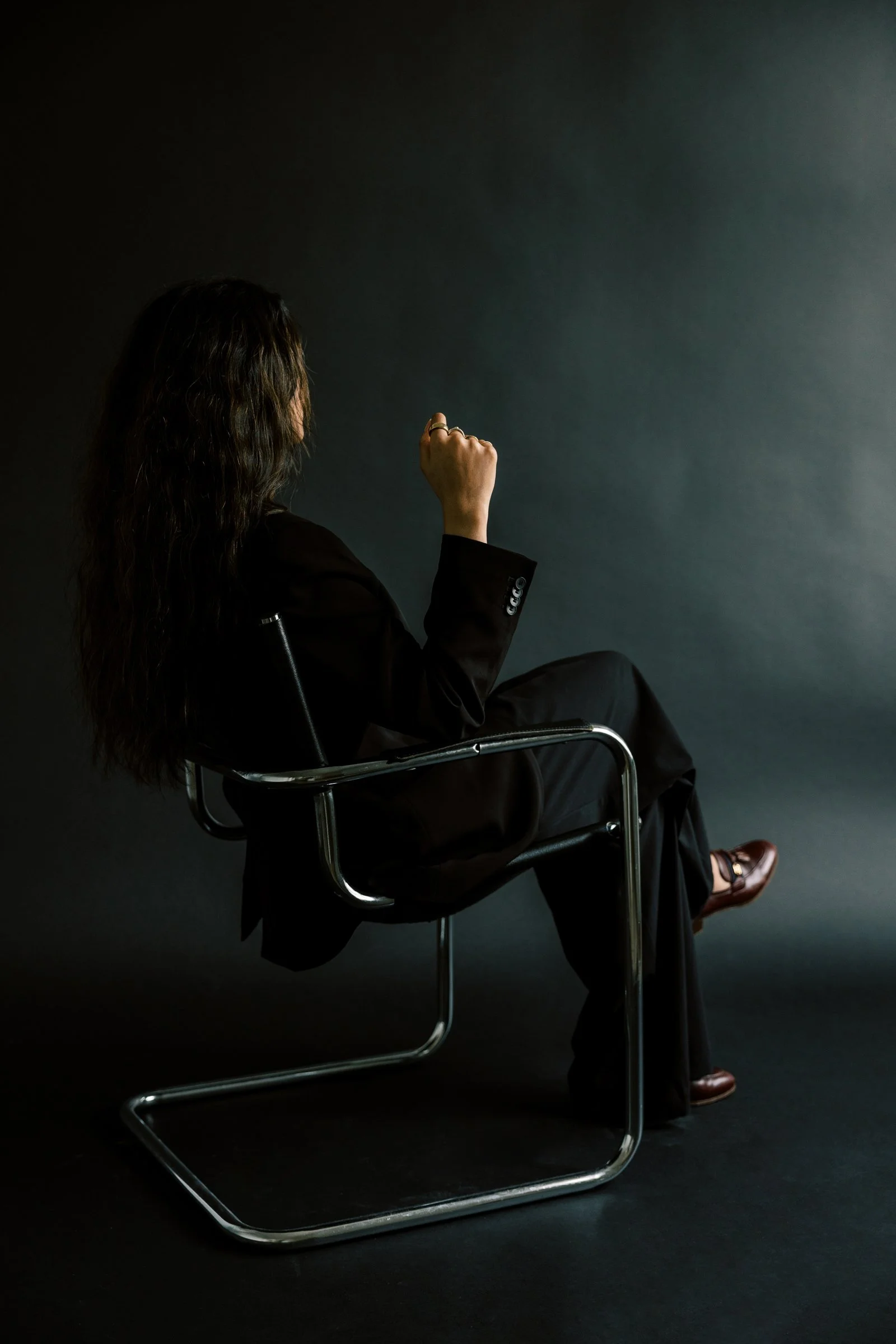 A woman with long dark hair, dressed in a black suit, sitting sideways on a modern metal chair against a dark background, with her left arm raised and her right hand resting on her lap.