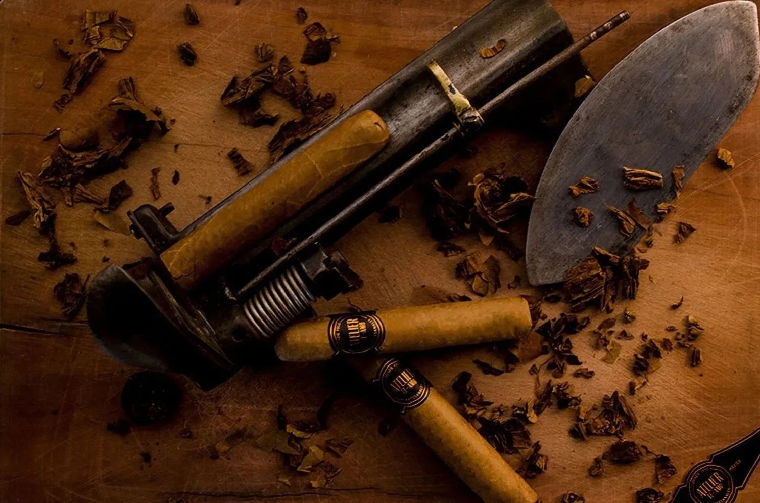 Still life of Atelier Te-Amo cigars on a weathered wooden table with a vintage tobacco cutter, torcedor's blade and scattered leaf trimmings.