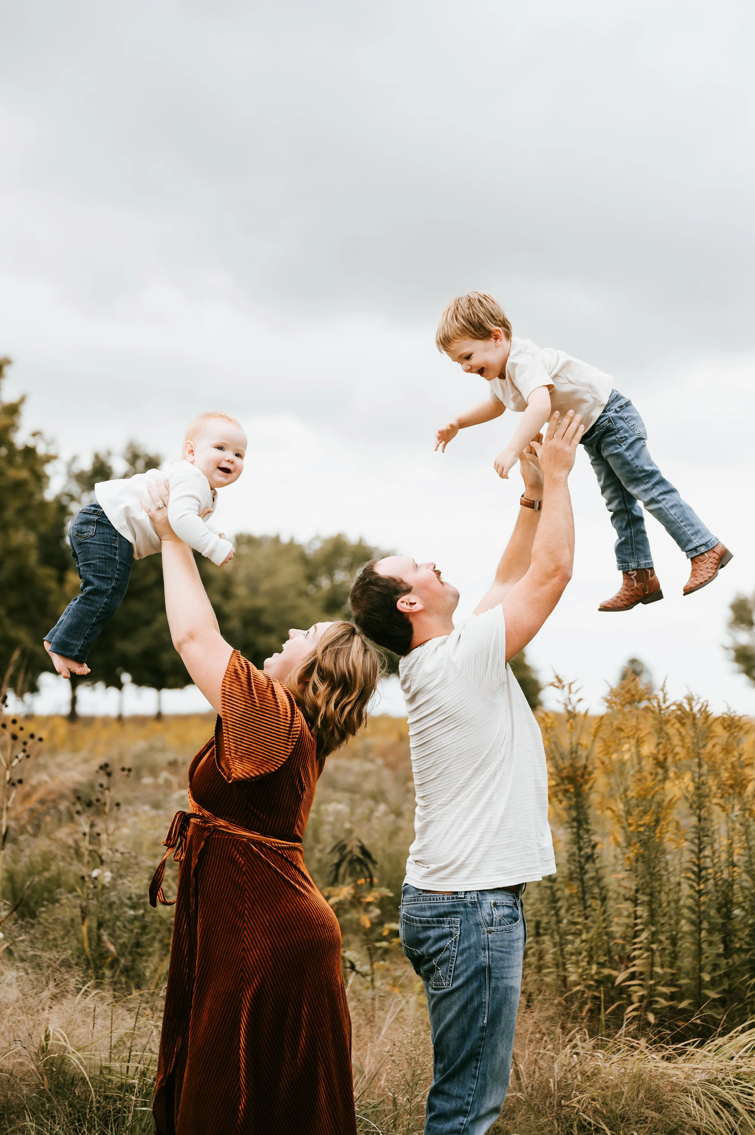 Family enjoying a playful moment outdoors: a man and woman lifting their two young children into the air in a field with trees in the background.