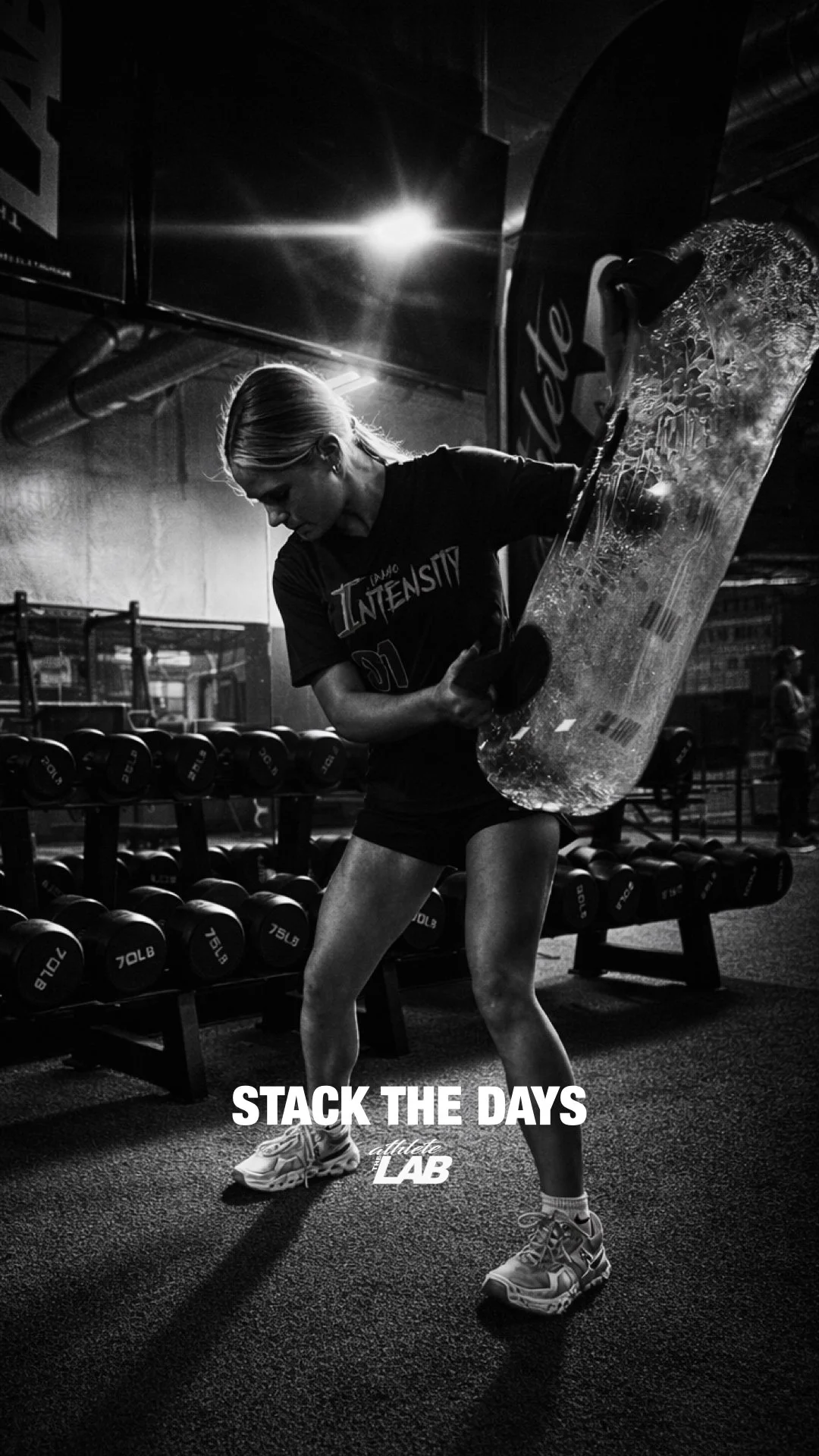 A woman lifting a large water tank in a gym with rack of dumbbells in the background. The photo is in black and white and has text that says 'Stack the Days' and 'athlete LAB'.