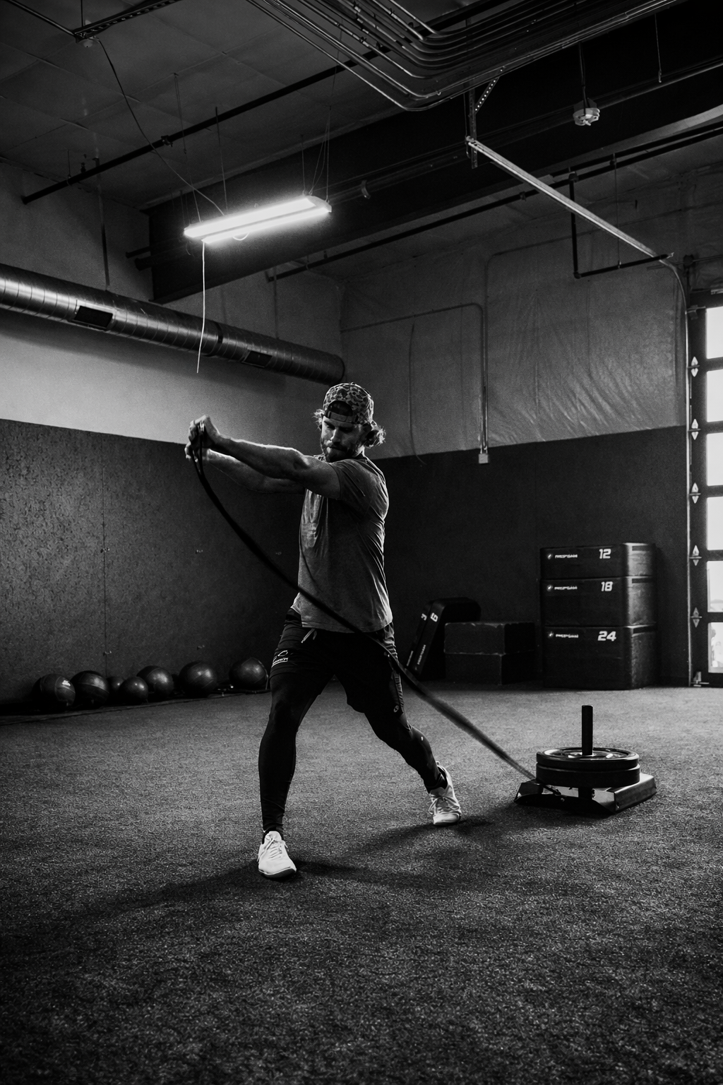 A man in a gym using a sled for strength training. He is pulling a weighted sled across the floor, wearing athletic clothing and a baseball cap.
