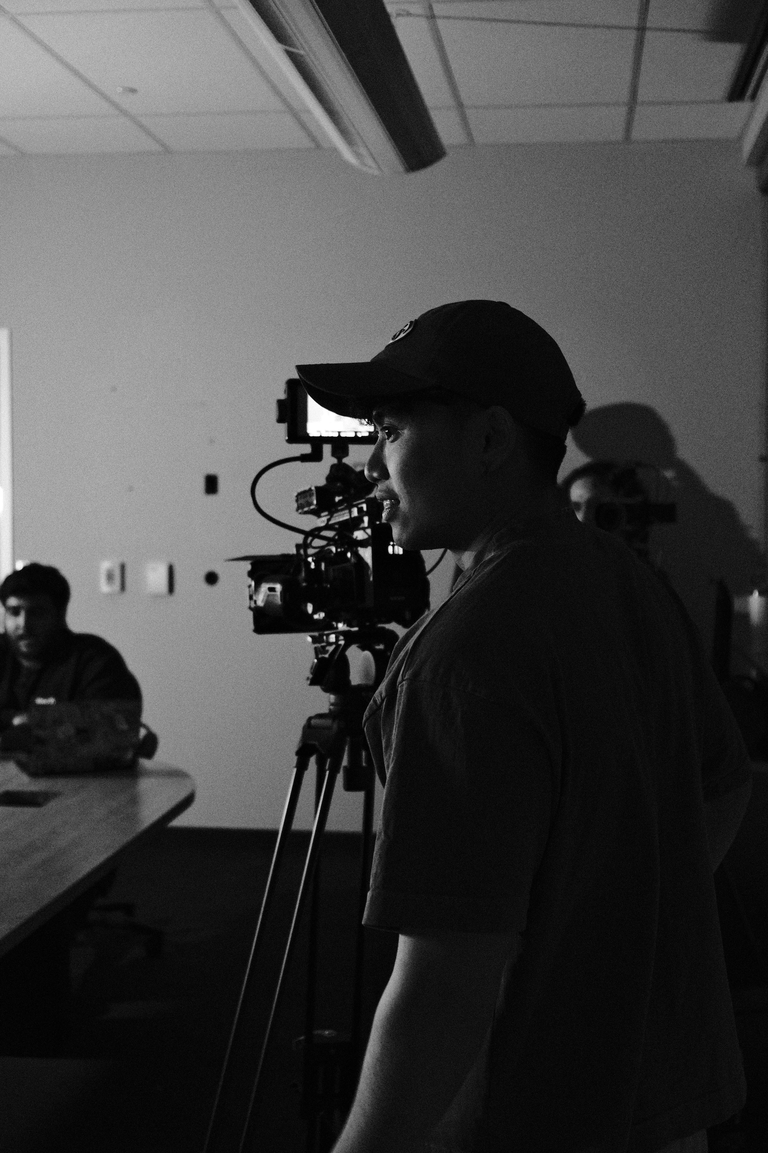 A person wearing a baseball cap operating a professional video camera in a dimly lit room, with a group of people sitting at a table in the background.