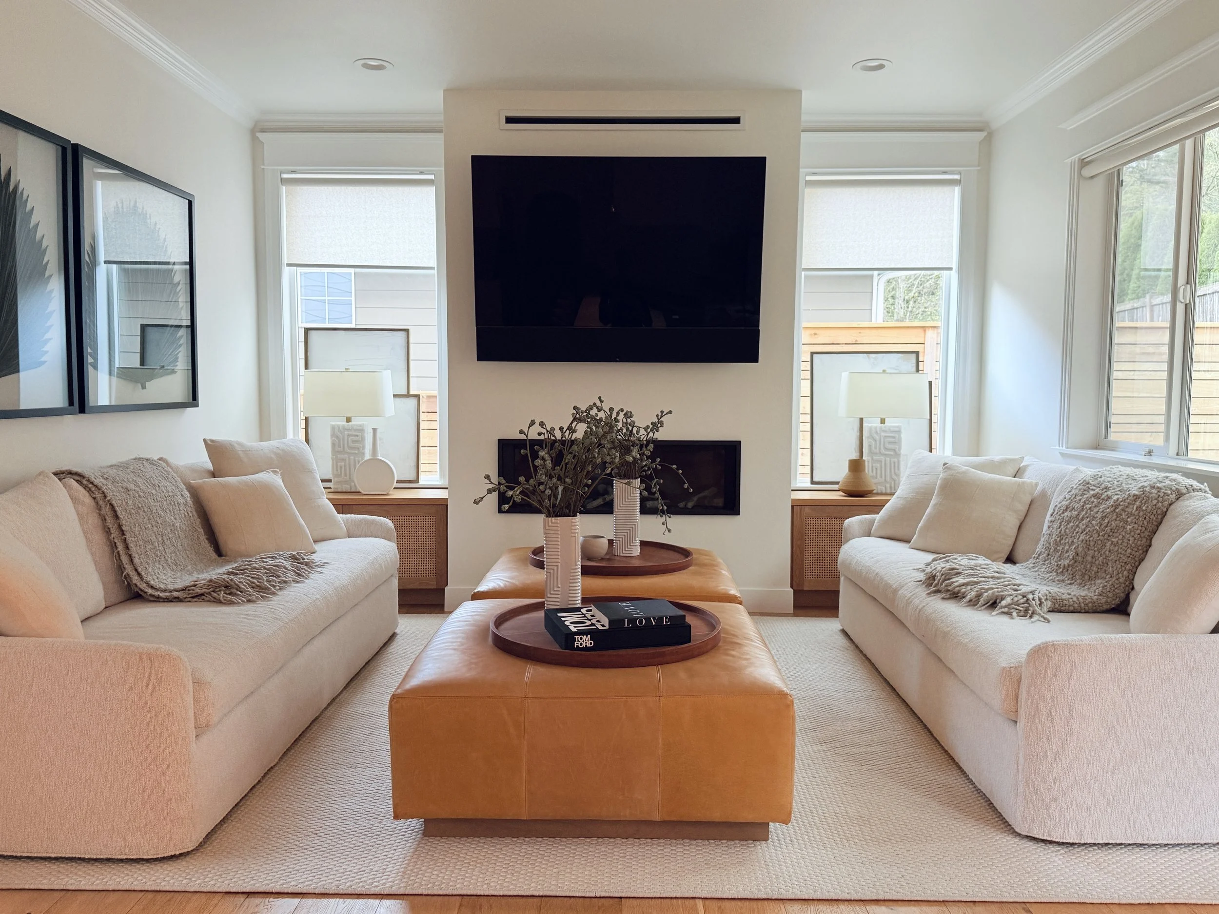 A symmetrical and serene living room with a cozy yet refined ambiance. Two cream-colored sofas face each other across a large tan leather ottoman that serves as a coffee table, topped with decorative trays and a vase of greenery. The space is grounde