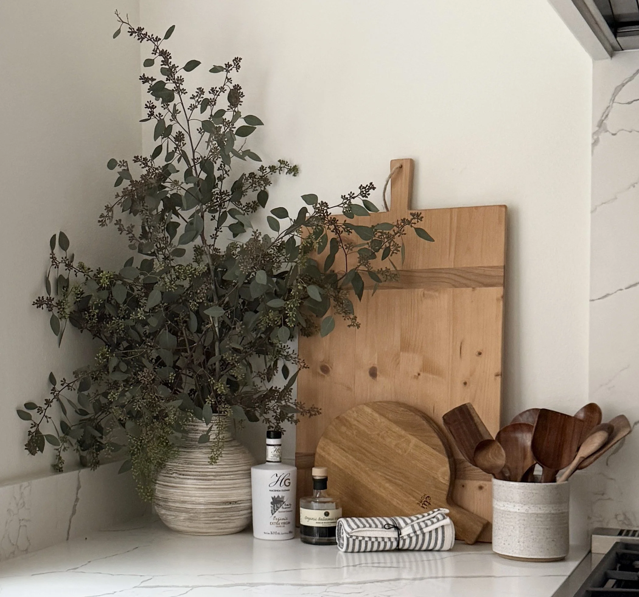 A beautifully styled kitchen countertop vignette with a natural, earthy aesthetic. The scene includes a large ceramic vase filled with eucalyptus branches, adding height and organic texture. Leaning against the wall are various wooden cutting boards 