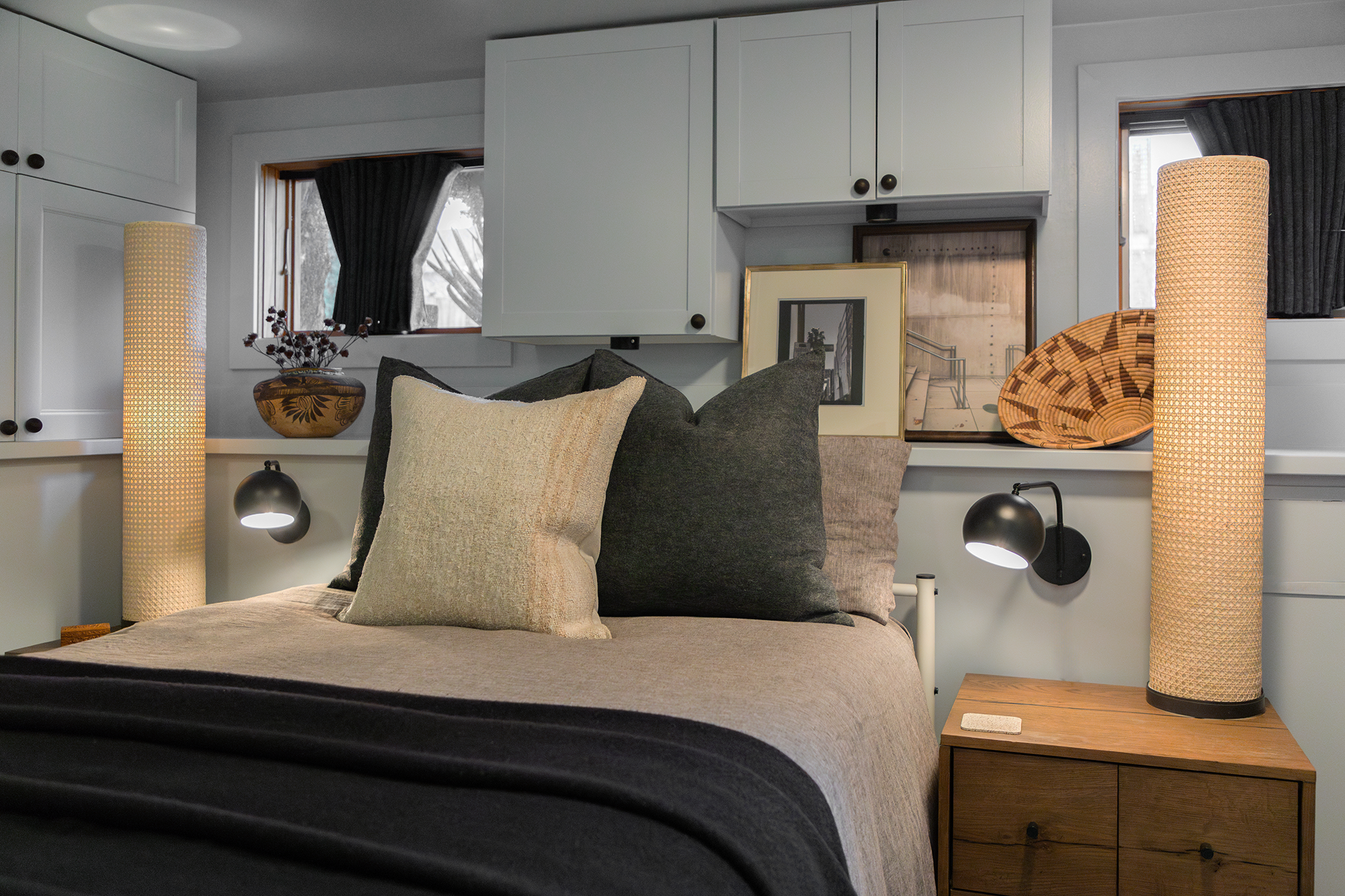 A compact yet stylish bedroom featuring a neatly made bed with layered neutral-toned linens and an assortment of soft pillows in shades of beige, gray, and charcoal. Flanking the bed are two cylindrical woven floor lamps that cast a warm, ambient glo