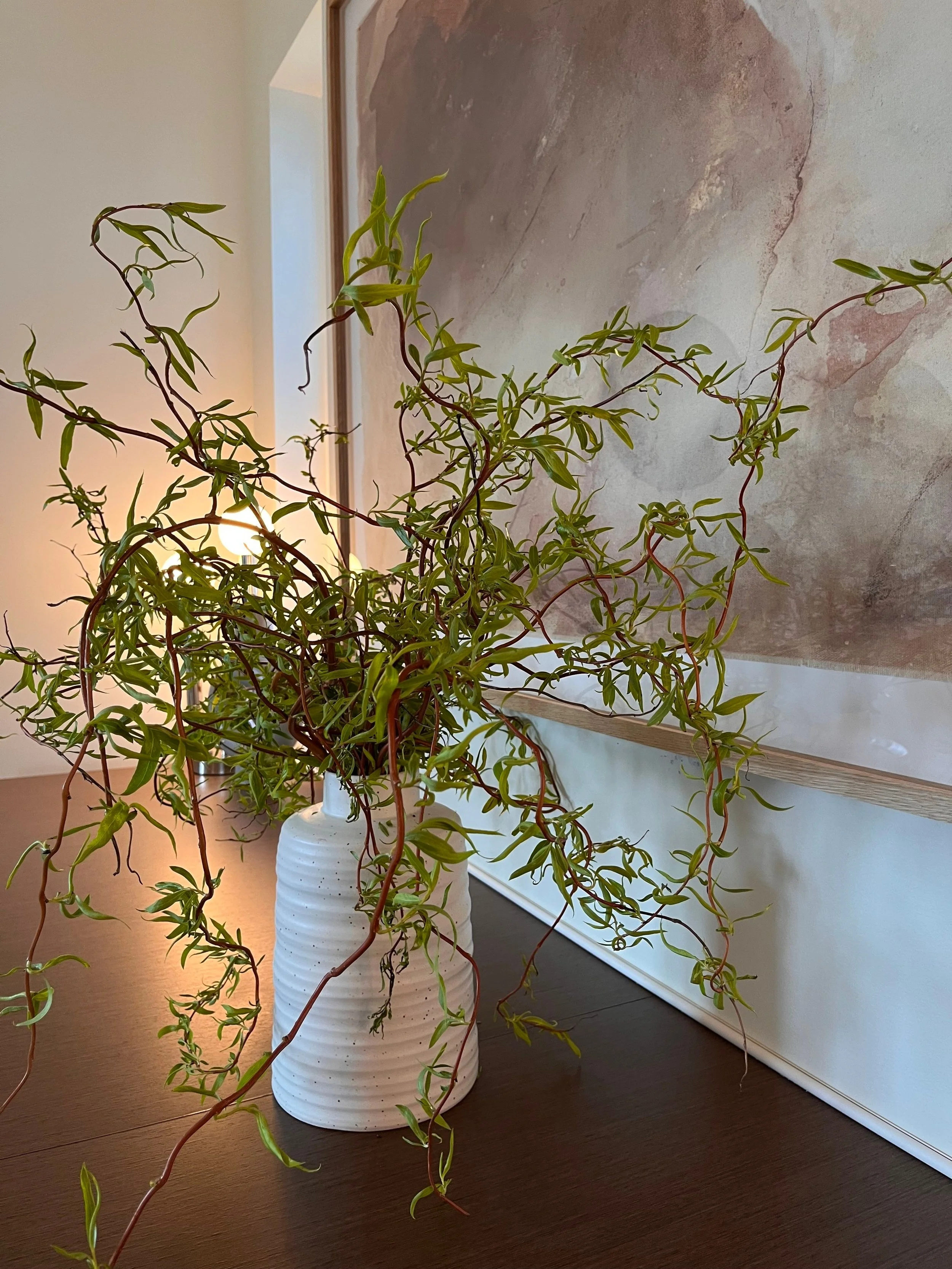 A close-up of a modern decorative arrangement featuring a sculptural, twisted green branch placed in a white ribbed ceramic vase. The vase rests on a dark wood surface, likely a console or sideboard. In the background, a large abstract artwork in sof