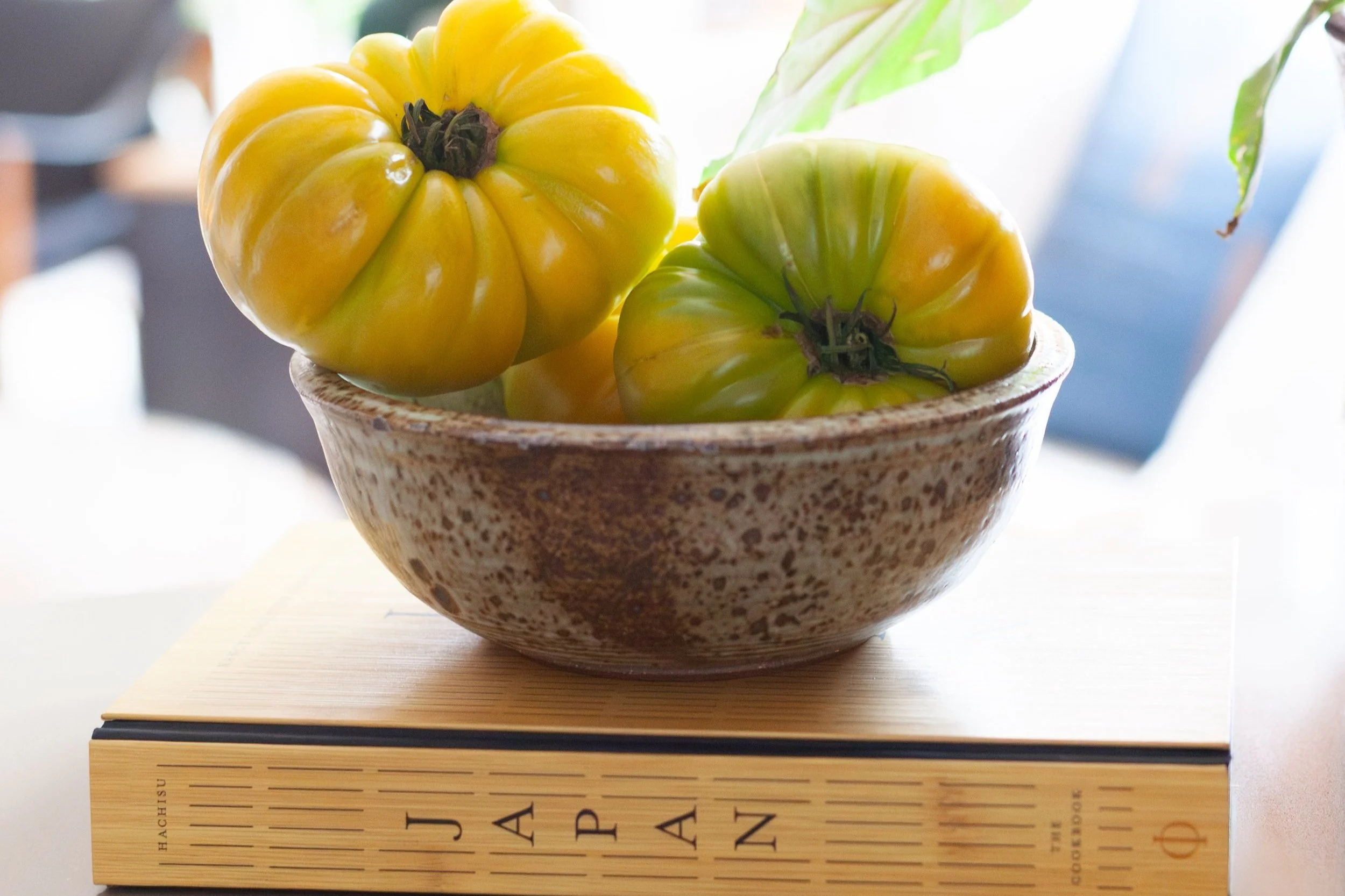 A close-up image of a rustic ceramic bowl filled with large, heirloom yellow-green tomatoes, resting on top of a hardcover book titled "Japan." Soft natural light illuminates the scene, highlighting the organic textures of the tomatoes and the earthy